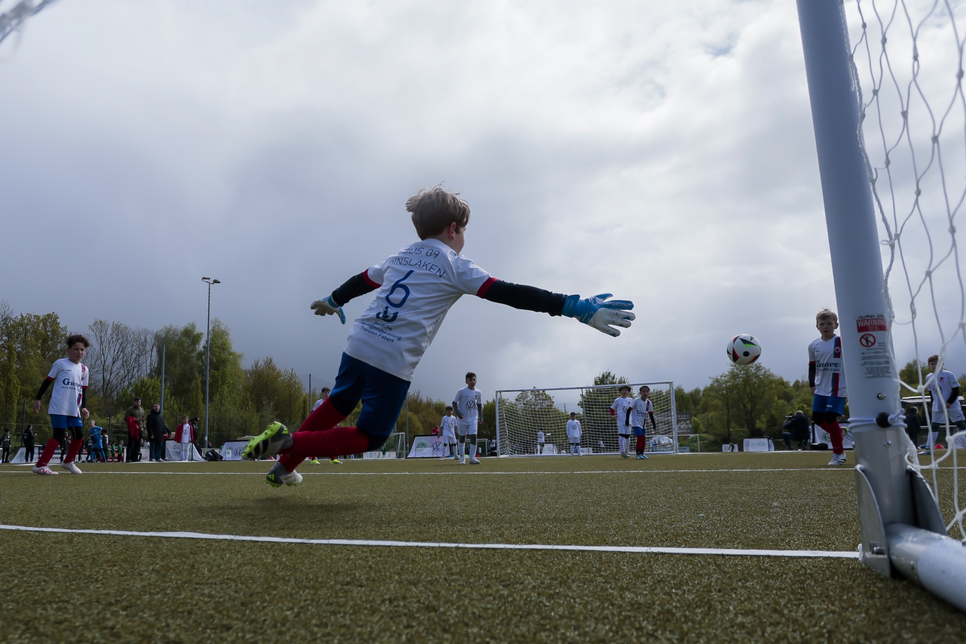 [{"clang_id":1,"value":"Junge springt als Torwart mit ausgestreckten Armen nach einem Fußball. Im Hintergrund weitere Spieler auf einem grünen Fußballfeld. Bewölkter Himmel."}]