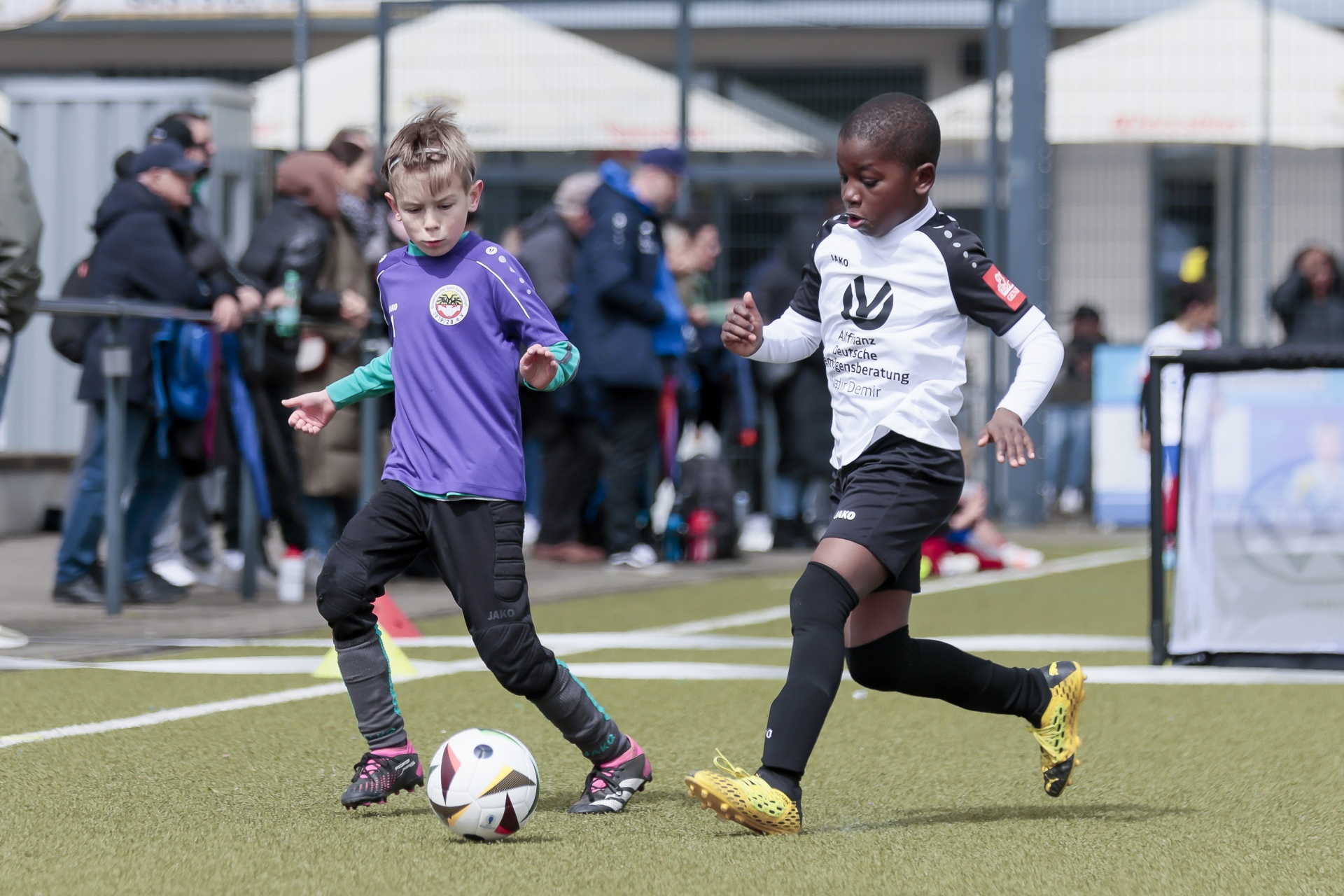 [{"clang_id":1,"value":"Zwei Fußballspieler, ein Junge mit dunkler Haut und ein Junge mit hellen Haaren, dribbeln auf einem Kunstrasenplatz. Im Hintergrund Zuschauer und ein Stadiondach."}]