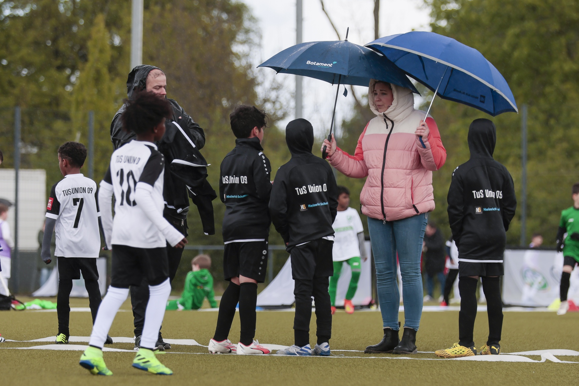 [{"clang_id":1,"value":"Fußballspieler mit Regenbekleidung stehen auf einem nassen Rasen. Eine Frau hält zwei Regenschirme über einige Kinder. Im Hintergrund Zuschauer."}]