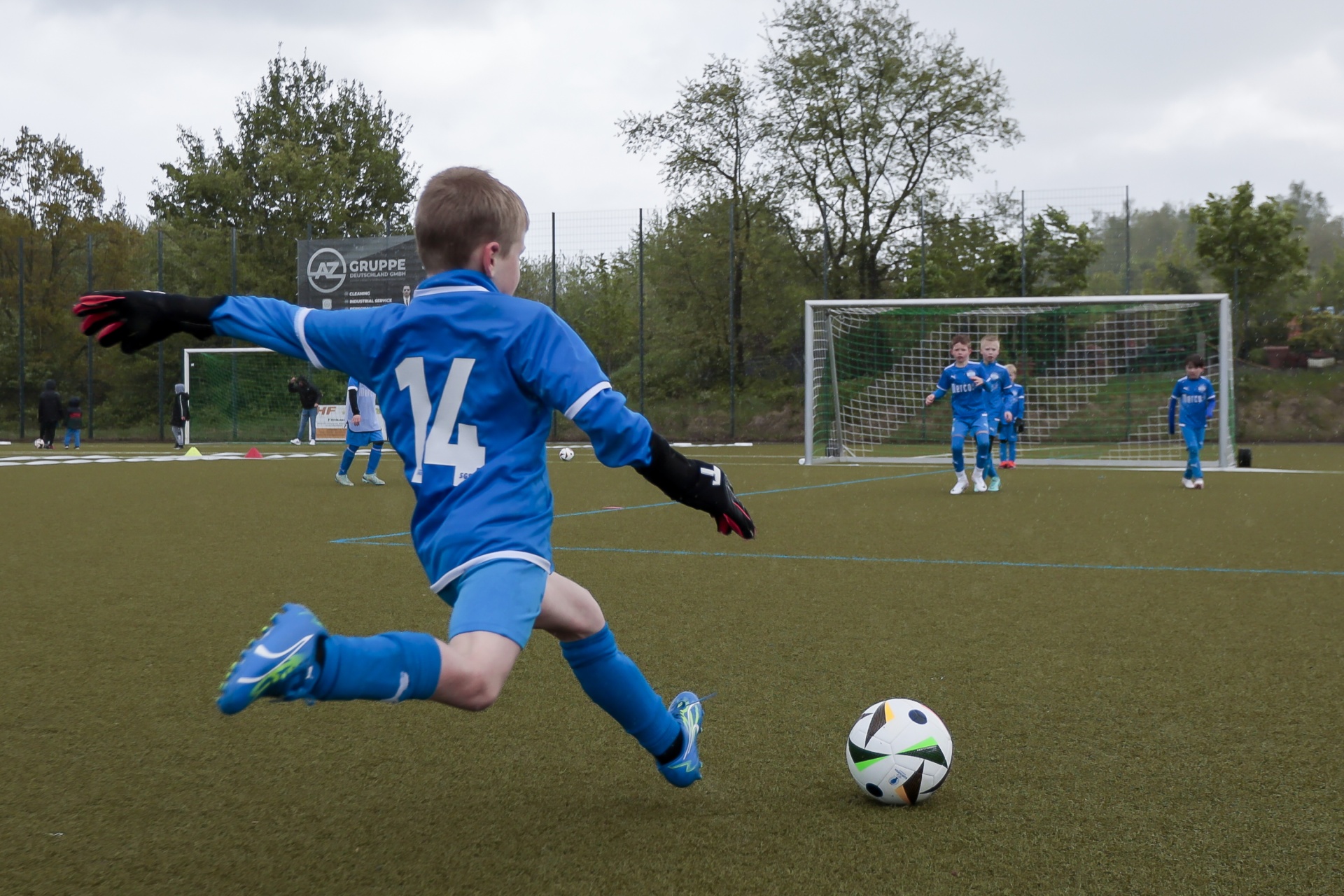 [{"clang_id":1,"value":"Junge kickt Fußball auf grünem Rasen. Im Hintergrund Fußballtor und weitere Spieler. Bewölkter Himmel, Bäume."}]