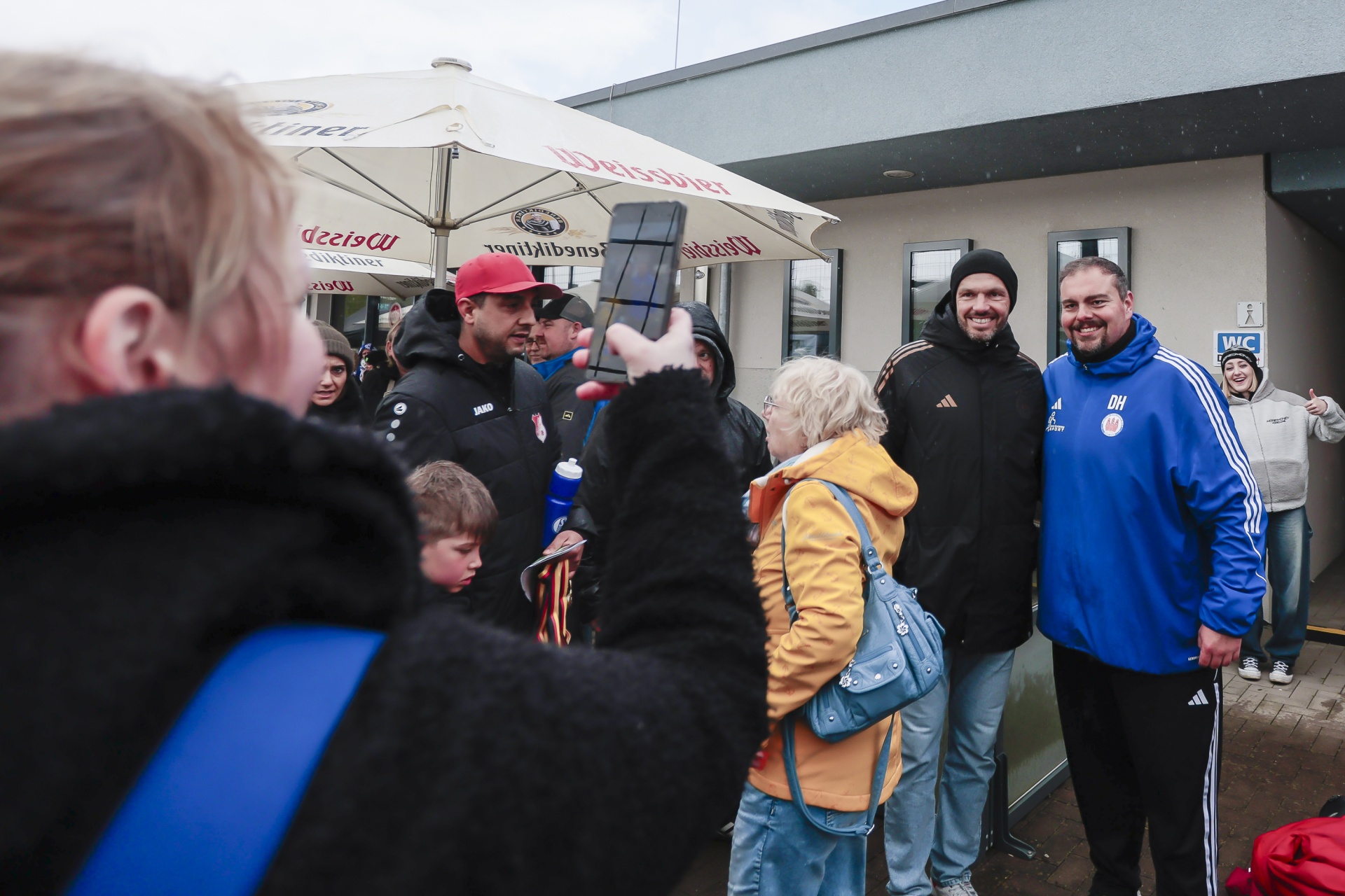 [{"clang_id":1,"value":"Menschengruppe vor Gebäude mit Sonnenschirmen. Im Vordergrund zwei lächelnde Männer in blauer Sportbekleidung, umgeben von Kindern und Erwachsenen."}]