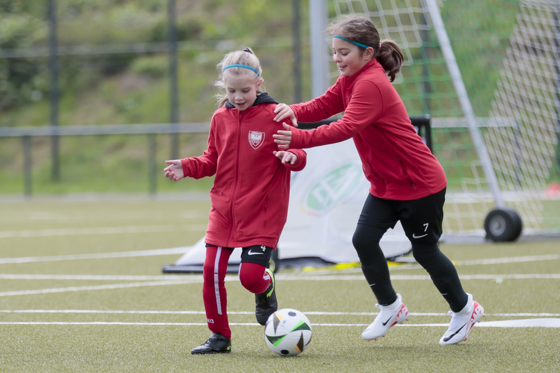 [{"clang_id":1,"value":"Zwei Mädchen trainieren Fußball auf einem Kunstrasenplatz. Ein Mädchen dribbelt den Ball, das andere begleitet sie und hält sie leicht am Arm."}]