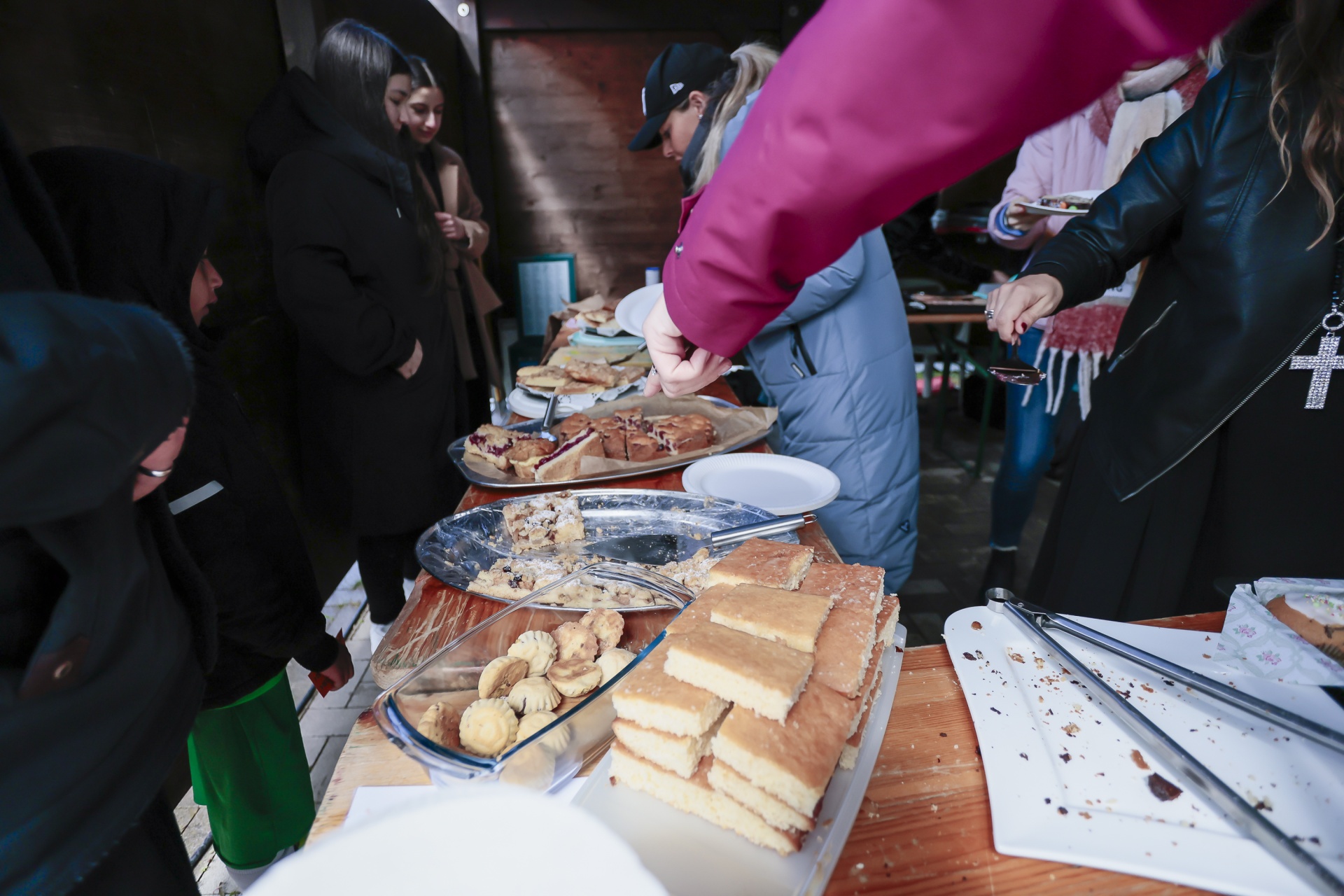[{"clang_id":1,"value":"Menschen stehen um einen Tisch mit verschiedenen Kuchen und Gebäck. Eine Frau mit pinkfarbener Pufferjacke greift nach einem Stück Kuchen."}]