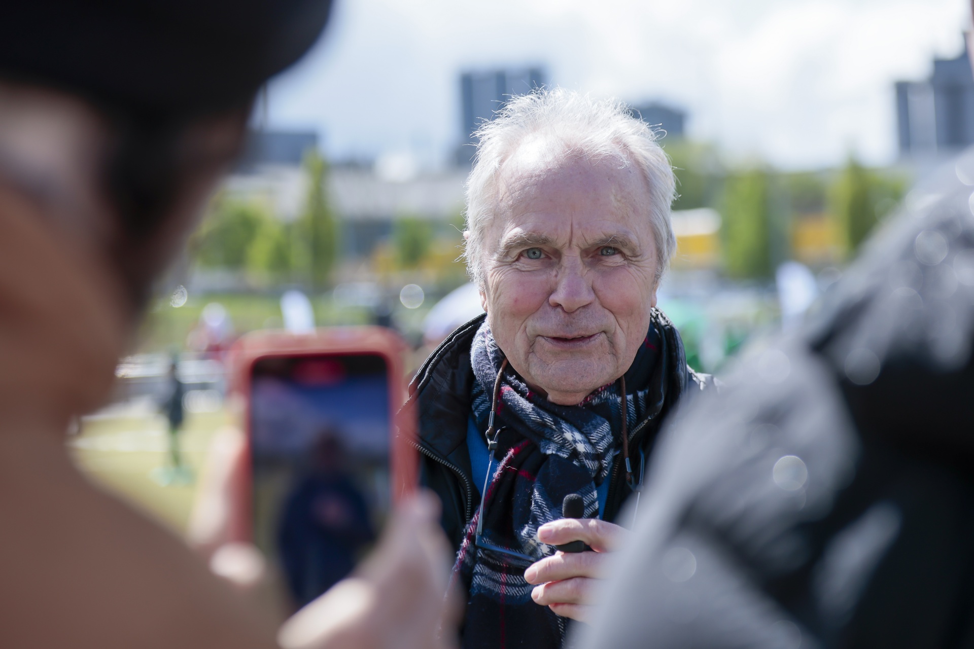 [{"clang_id":1,"value":"Älterer Mann mit weißen Haaren und Schal wird von Personen mit Smartphones gefilmt. Hintergrund ist eine grüne Wiese und Gebäude."}]