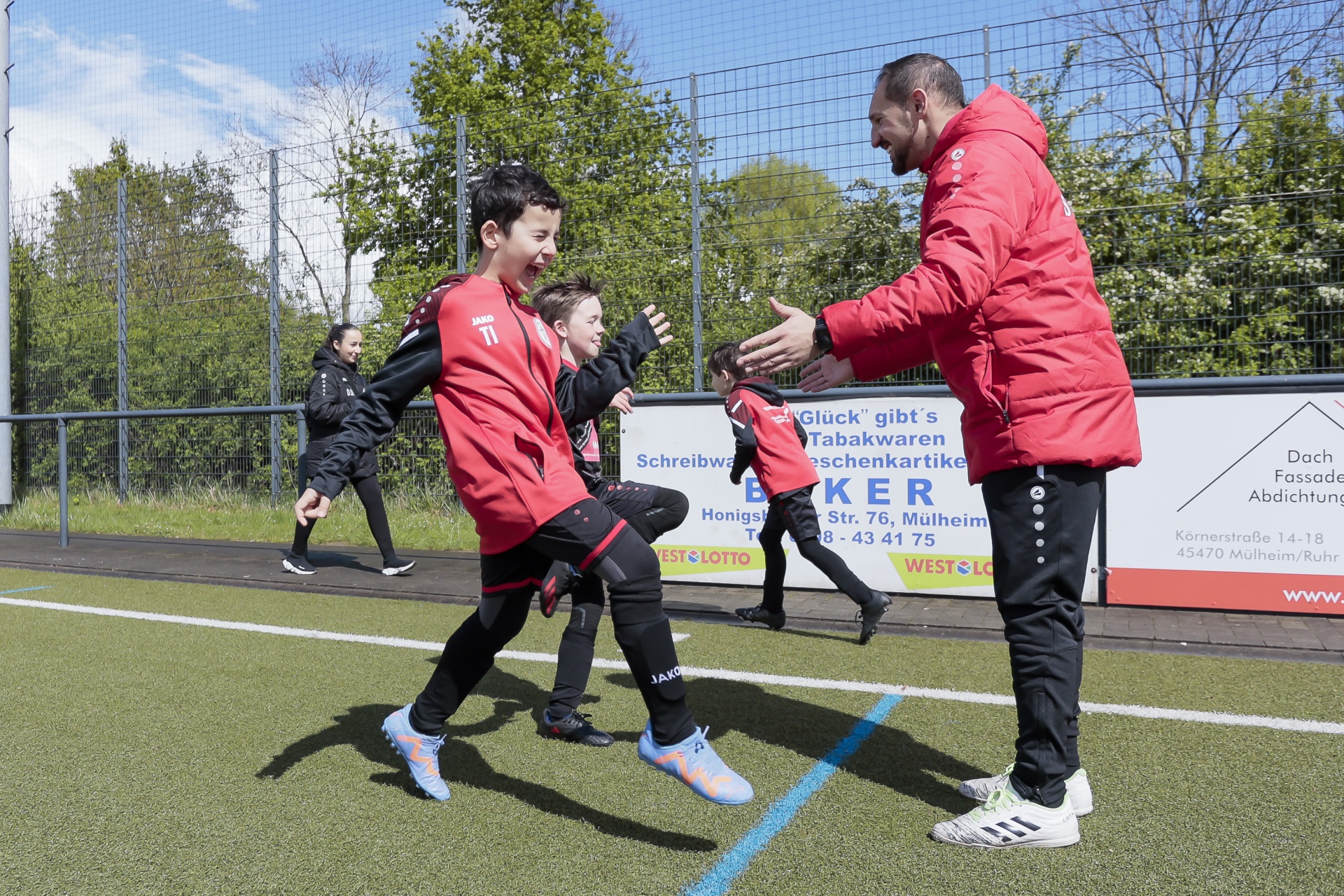 [{"clang_id":1,"value":"Fußballtrainer in roter Jacke unterstützt zwei junge Fußballspieler beim Dehnen auf einem grünen Kunstrasenplatz. Im Hintergrund ein Zaun und Werbebanner."}]