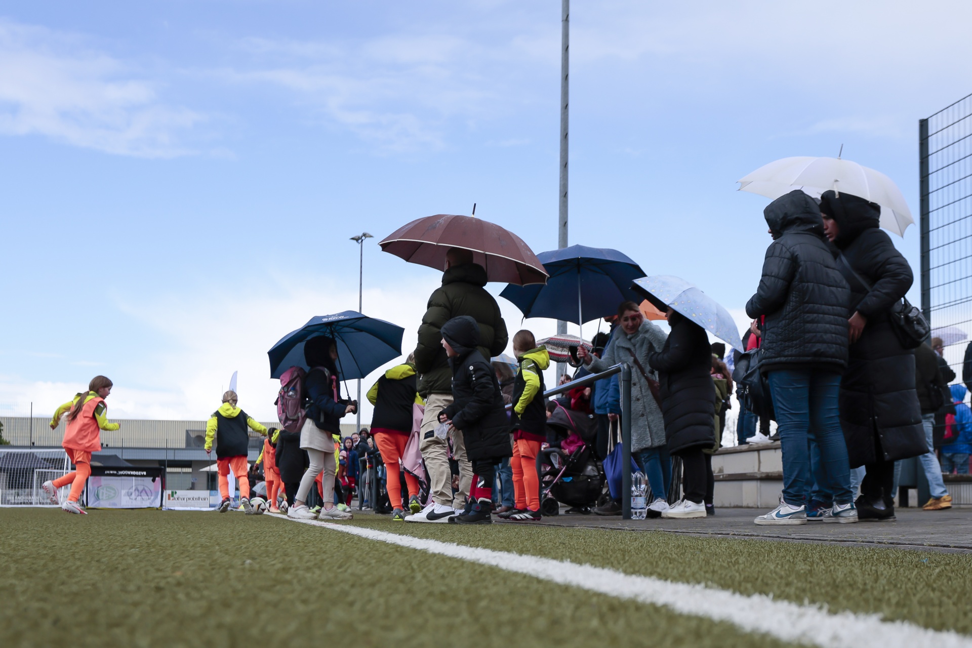 [{"clang_id":1,"value":"Zuschauer mit Regenschirmen stehen am Rand eines grünen Sportplatzes. Im Hintergrund ein Stadion und ein blauer Himmel mit Wolken."}]
