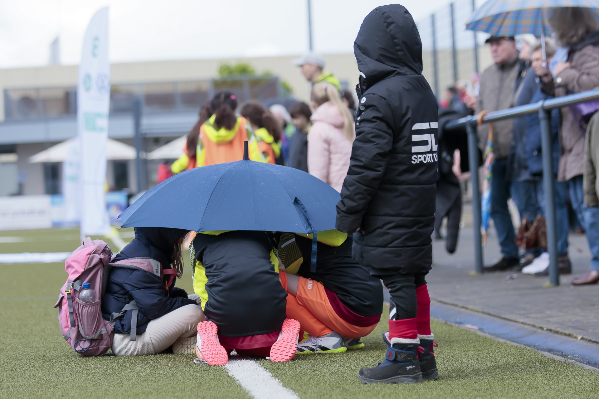 [{"clang_id":1,"value":"Kind mit Kapuzenjacke steht neben zwei Personen unter einem Regenschirm. Im Hintergrund Menschen und ein Gebäude mit Vordach, vermutlich bei Regen."}]