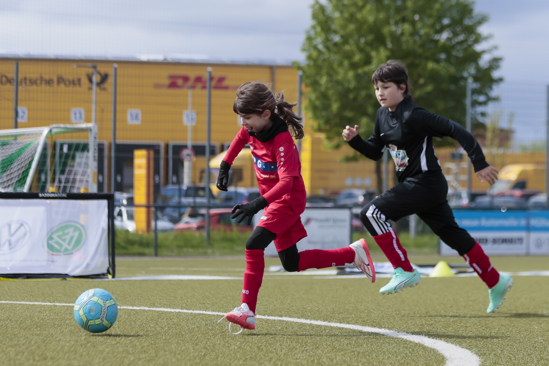 [{"clang_id":1,"value":"Zwei Kinder spielen Fußball auf einem Kunstrasenplatz. Ein Mädchen in Rot schießt auf den Ball, ein Junge in Schwarz rennt daneben. Im Hintergrund Zaun und Werbung."}]