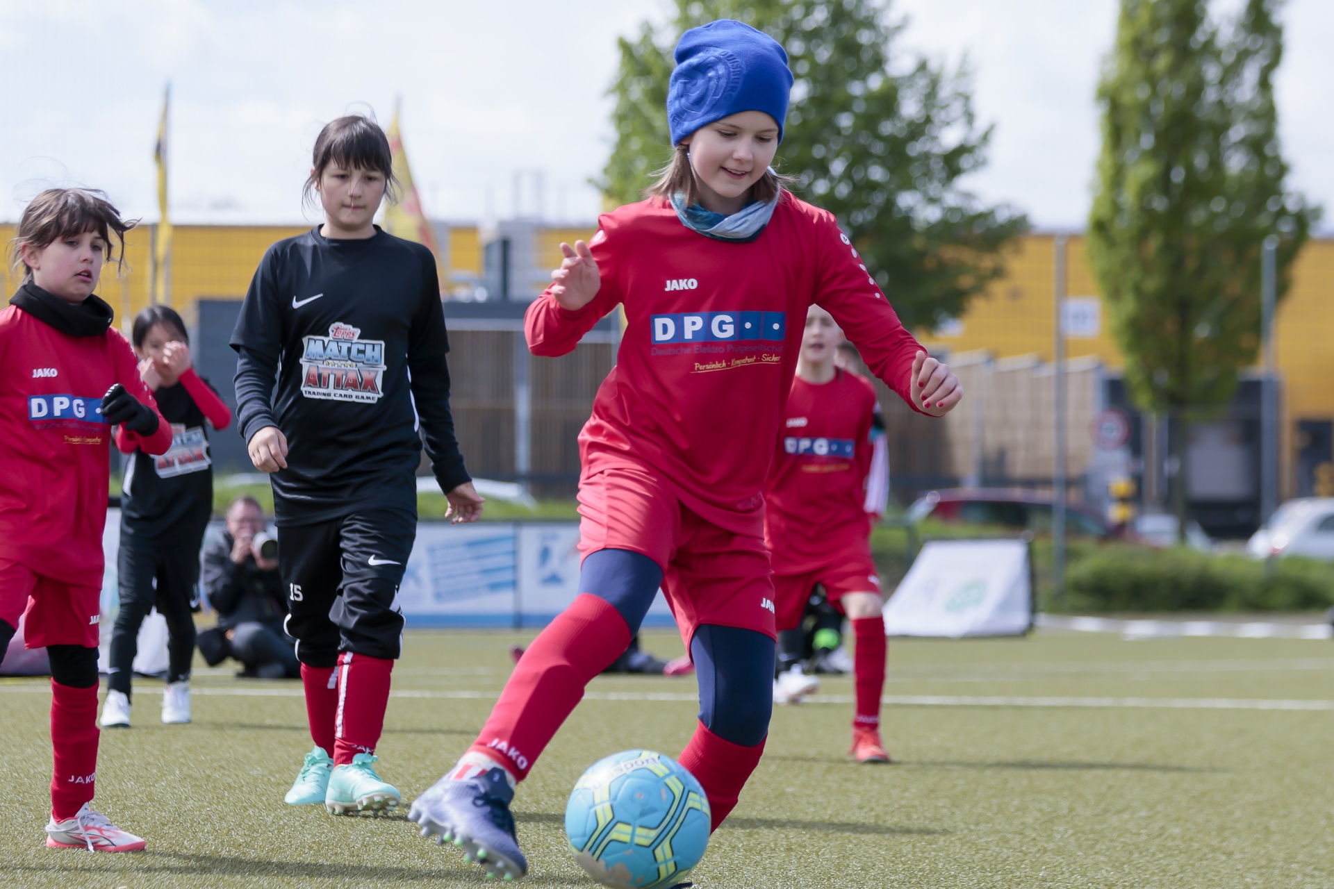 [{"clang_id":1,"value":"Fußballspiel. Ein Kind in rotem Trikot dribbelt den Ball auf einem grünen Kunstrasenplatz. Im Hintergrund weitere Kinder und ein Gebäudekomplex."}]