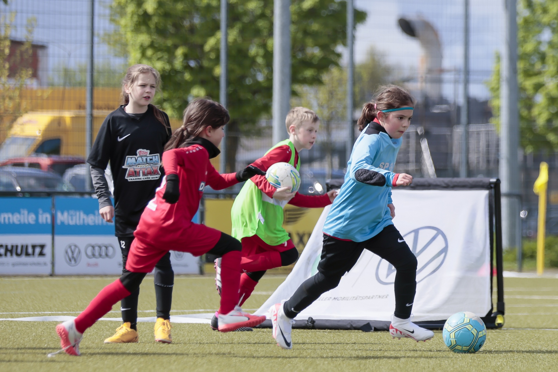 [{"clang_id":1,"value":"Fußballspiel: Vier Kinder auf grünem Kunstrasen, zwei Mädchen rennen dem Ball entgegen, ein Junge hält ihn, im Hintergrund Tor und Werbung."}]