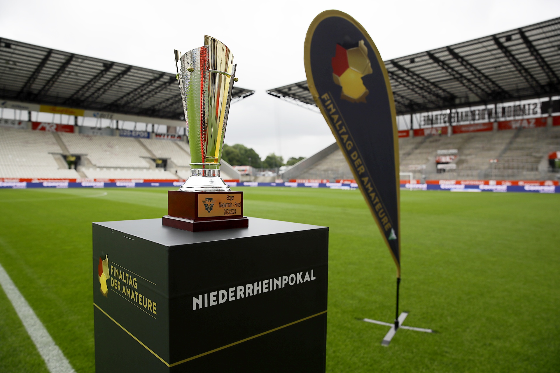 [{"clang_id":1,"value":"Fußballstadion: Pokal auf Podest im Vordergrund. Im Hintergrund leere Zuschauertribünen und Rasenplatz. Banner mit Schriftzug \"Finaltag der Amateure\"."}]
