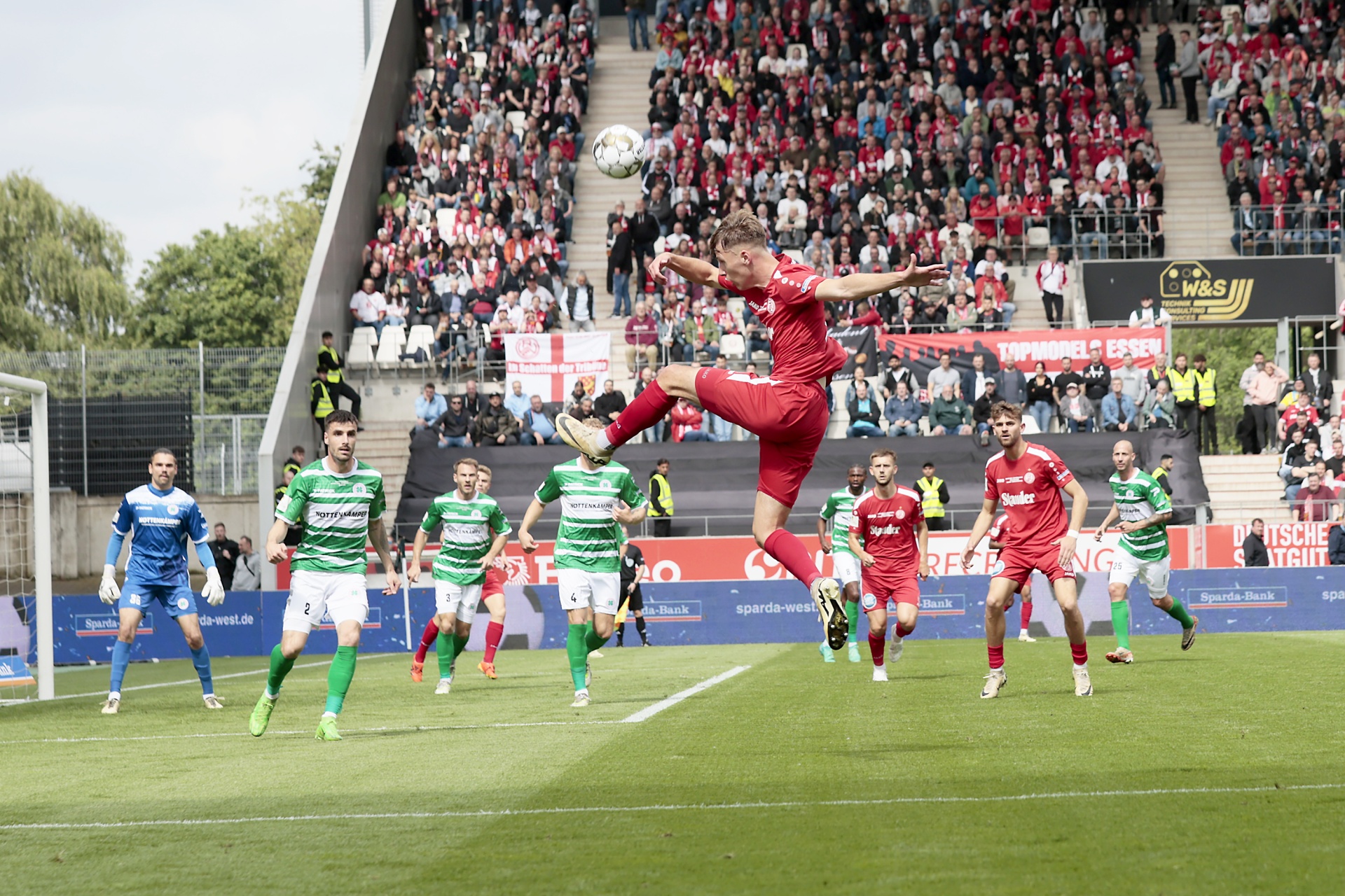 [{"clang_id":1,"value":"Fußballspiel: Spieler in rot springt und schießt Ball. Im Hintergrund dichtes Publikum auf roten Sitzen, weitere Spieler in grün und rot auf grünem Rasen."}]