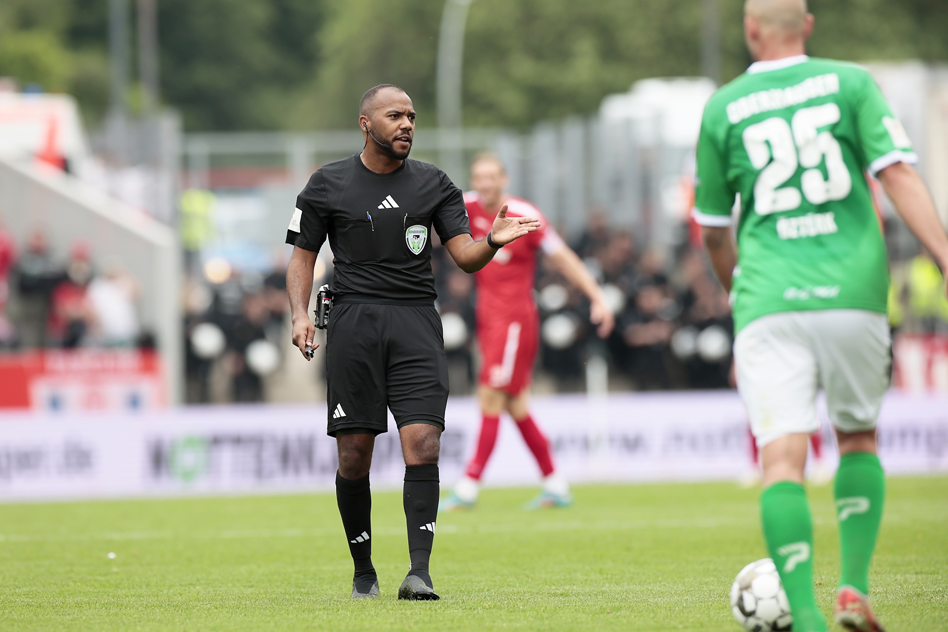 [{"clang_id":1,"value":"Fußballspiel: Schiedsrichter in schwarzer Uniform gestikuliert, Spieler mit Nummer 25 im Hintergrund. Rasenplatz und Zuschauerreihen erkennbar."}]