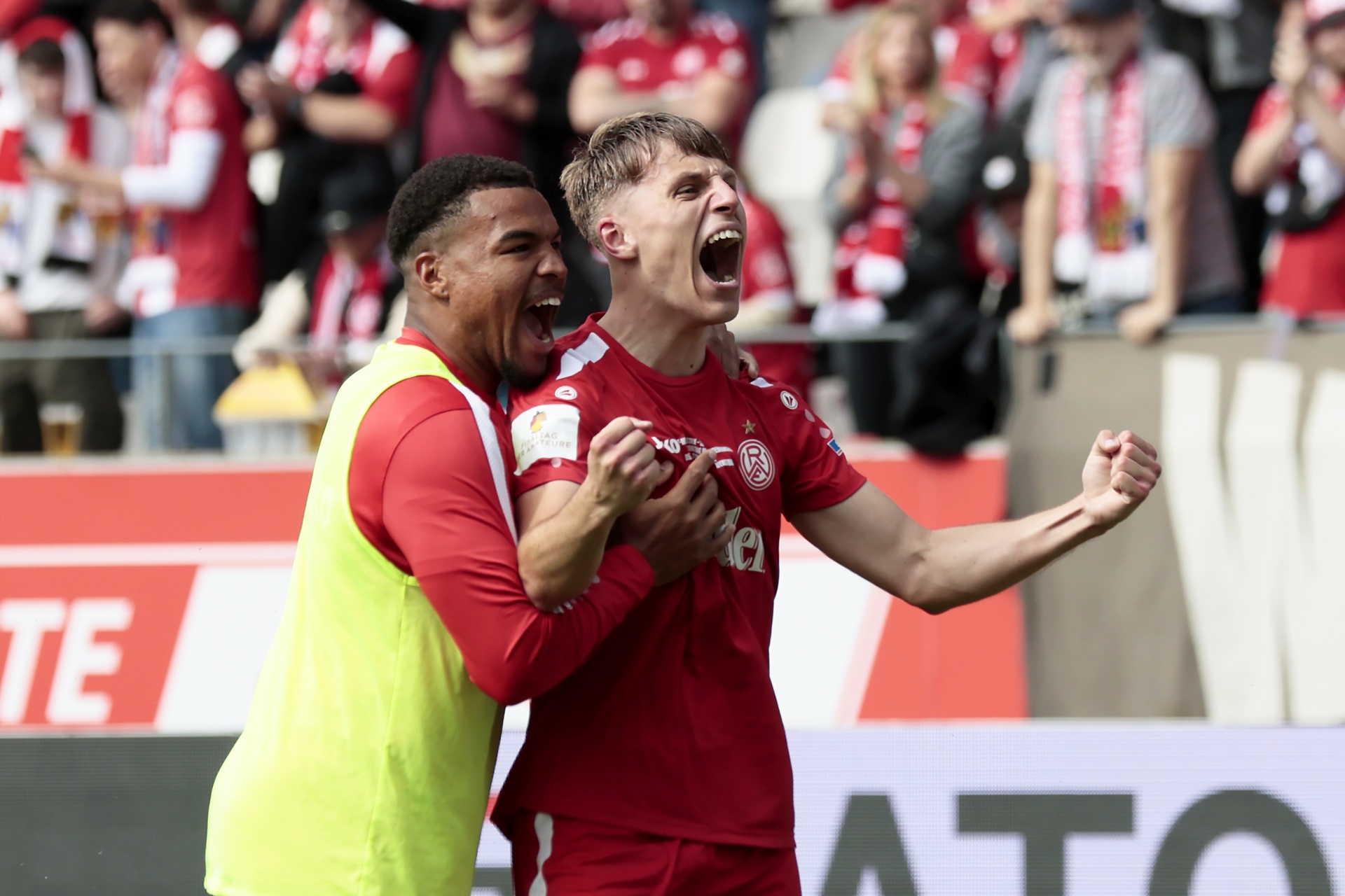 [{"clang_id":1,"value":"Zwei Fußballspieler, einer mit gelber Weste, jubeln vor einer gefüllten Tribüne mit rot gekleideten Fans. Im Hintergrund sind Werbebanner erkennbar."}]