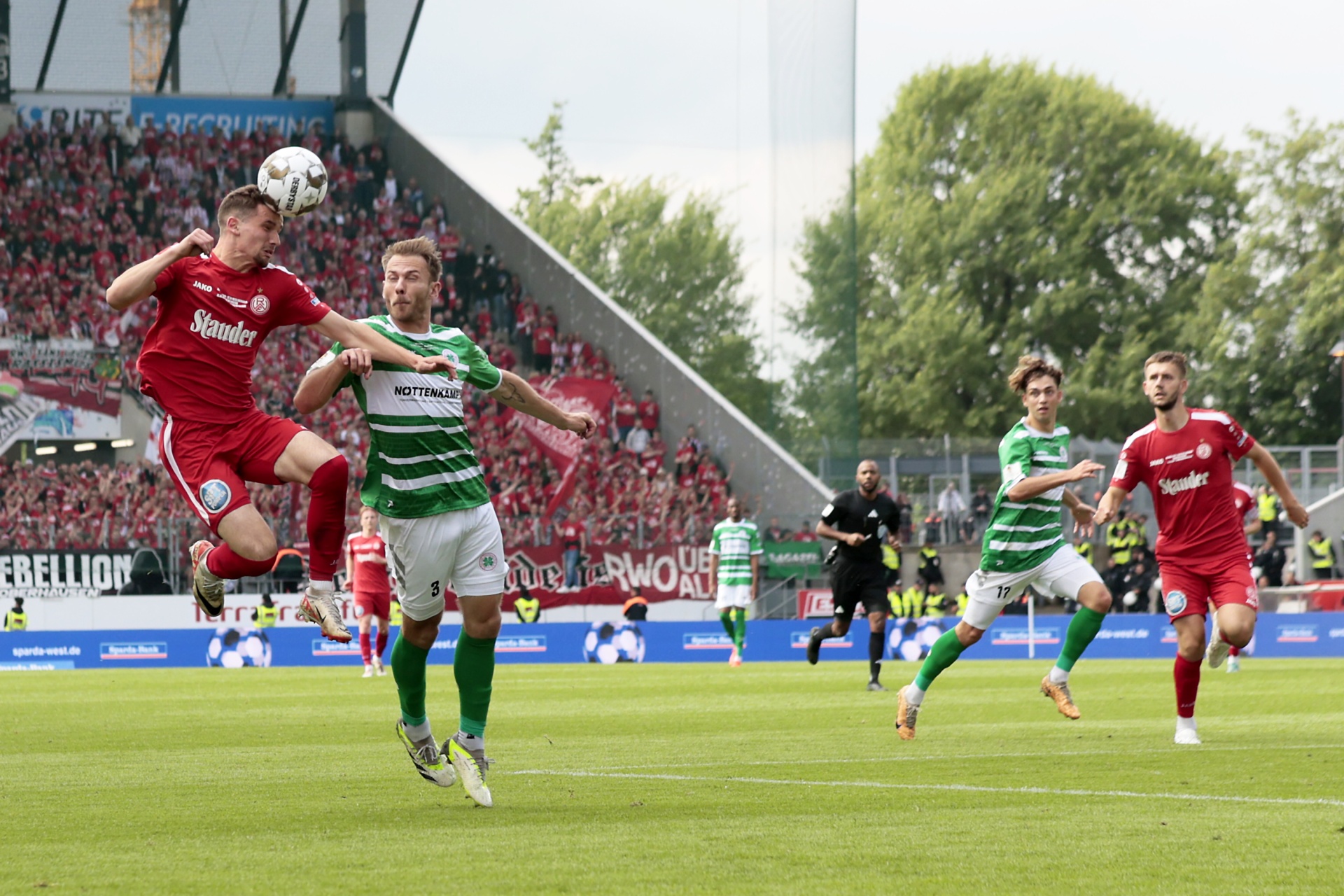[{"clang_id":1,"value":"Zwei Fußballspieler springen im Stadion um einen Ball. Im Hintergrund weitere Spieler und Zuschauer auf den Tribünen. Rasenplatz."}]