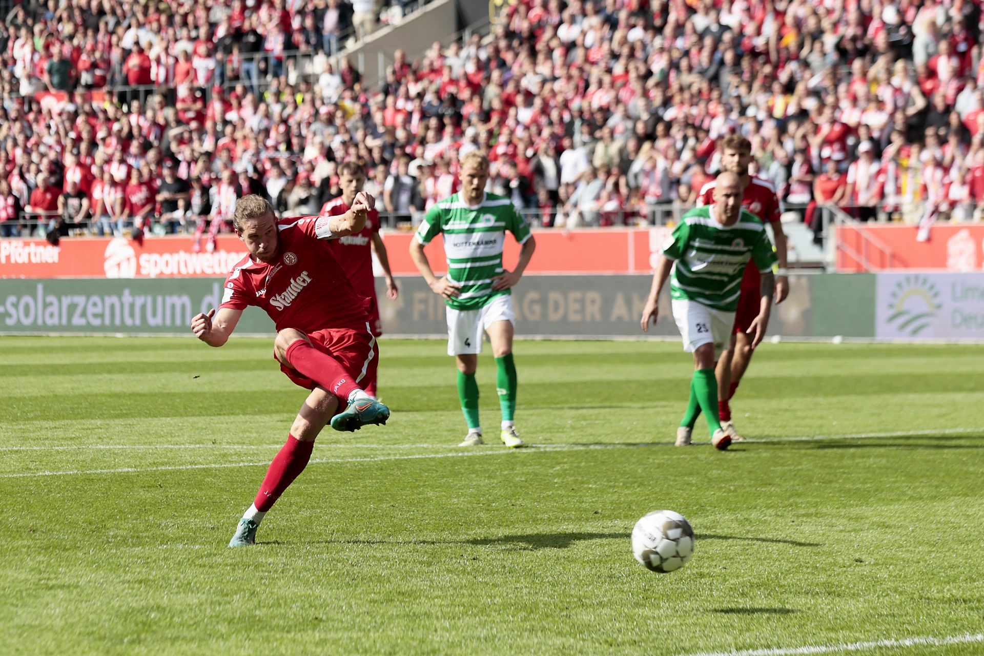 [{"clang_id":1,"value":"Fußballspiel: Spieler in roten Trikots schießt auf das Tor. Zwei Gegenspieler in grünen Trikots verteidigen. Im Hintergrund dichtes Publikum."}]