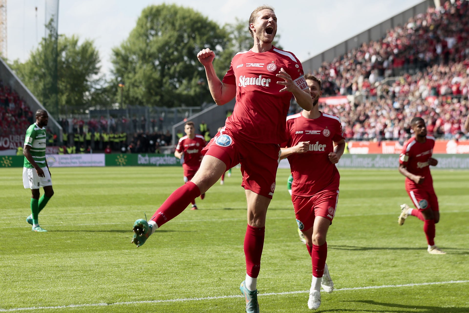 [{"clang_id":1,"value":"Fußballspieler in roten Trikots jubeln auf grünem Rasen, im Hintergrund Zuschauer und Stadion. Gegenüber stehen Spieler in grünen Trikots."}]