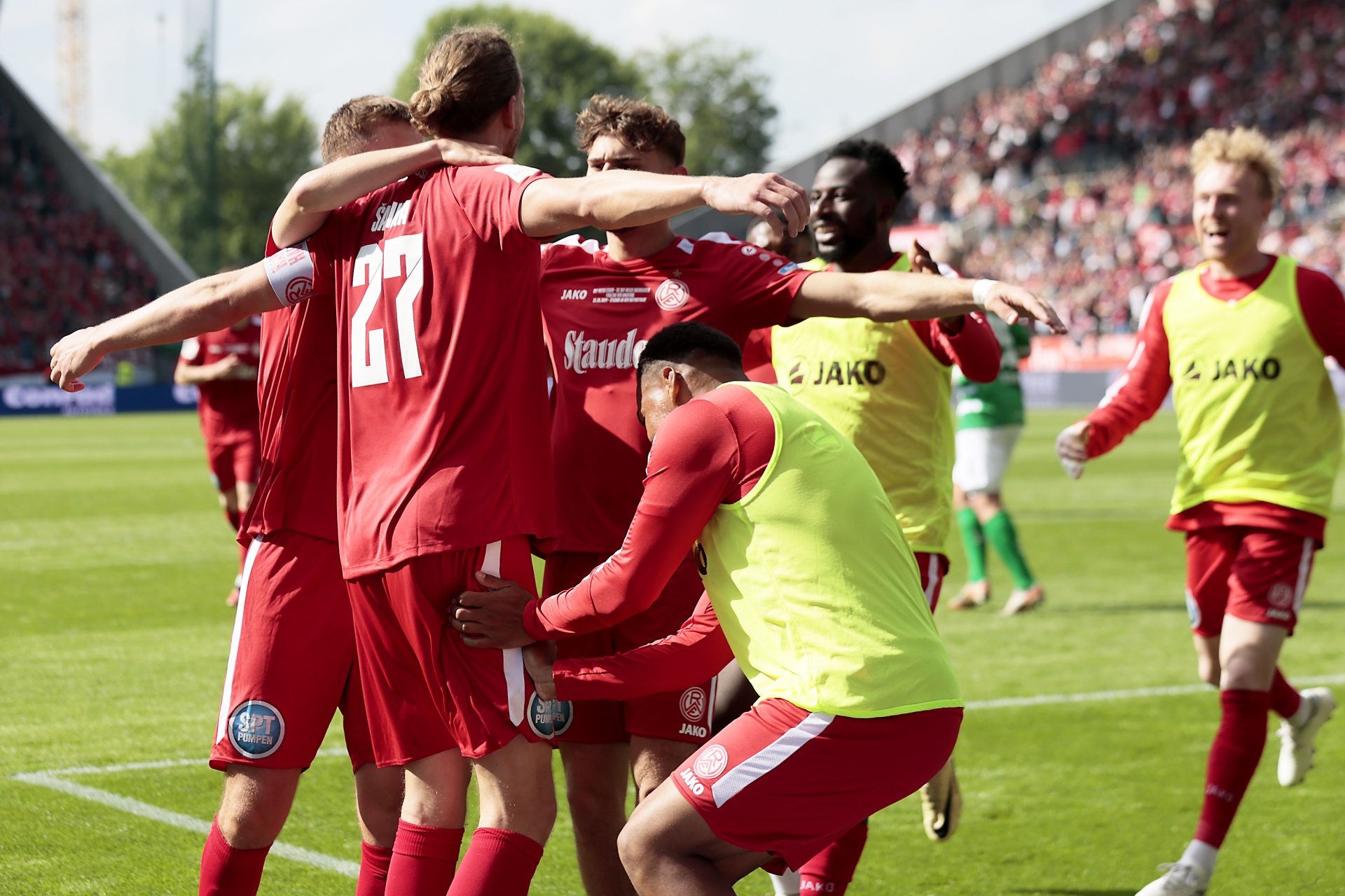 [{"clang_id":1,"value":"Fußballspieler in roten Trikots und Shorts feiern mit erhobenen Armen auf dem grünen Rasen. Einige tragen gelbe Trainingswesten. Im Hintergrund Zuschauer."}]