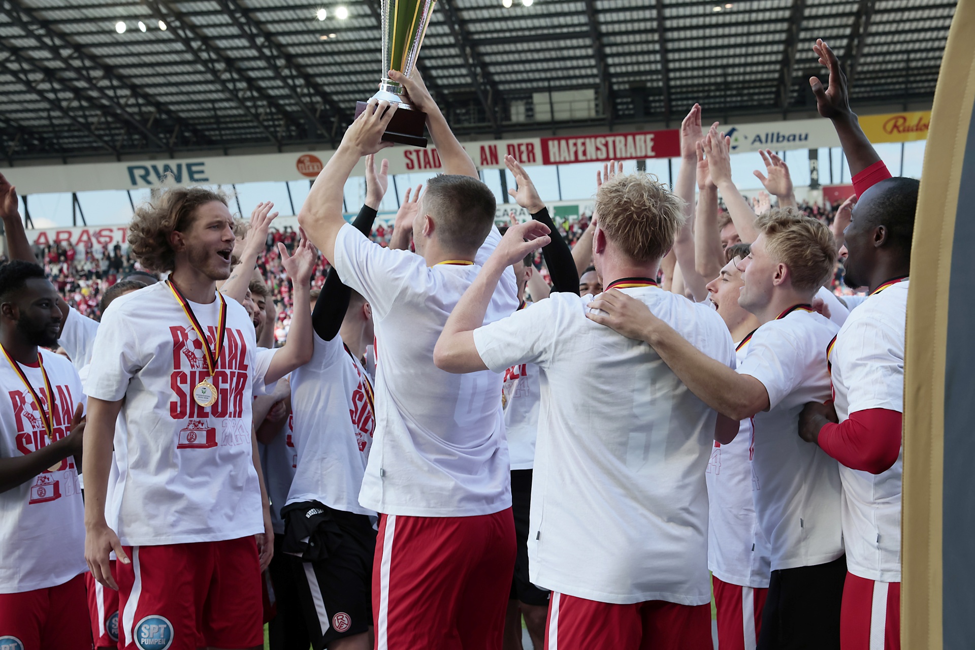 [{"clang_id":1,"value":"Fußballmannschaft feiert Sieg. Spieler mit weiß-roten Trikots und Pokal in der Hand, jubelnde Fans im Hintergrund. Stadion."}]