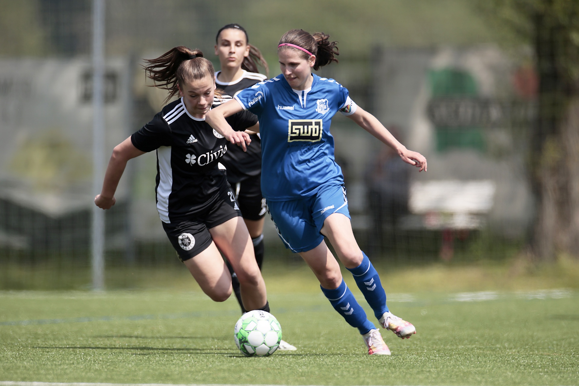 [{"clang_id":1,"value":"Zwei Fußballspielerinnen im Wettkampf. Links trägt eine Spielerin ein schwarz-weißes Trikot, rechts ein blaues. Im Hintergrund eine weitere Spielerin."}]
