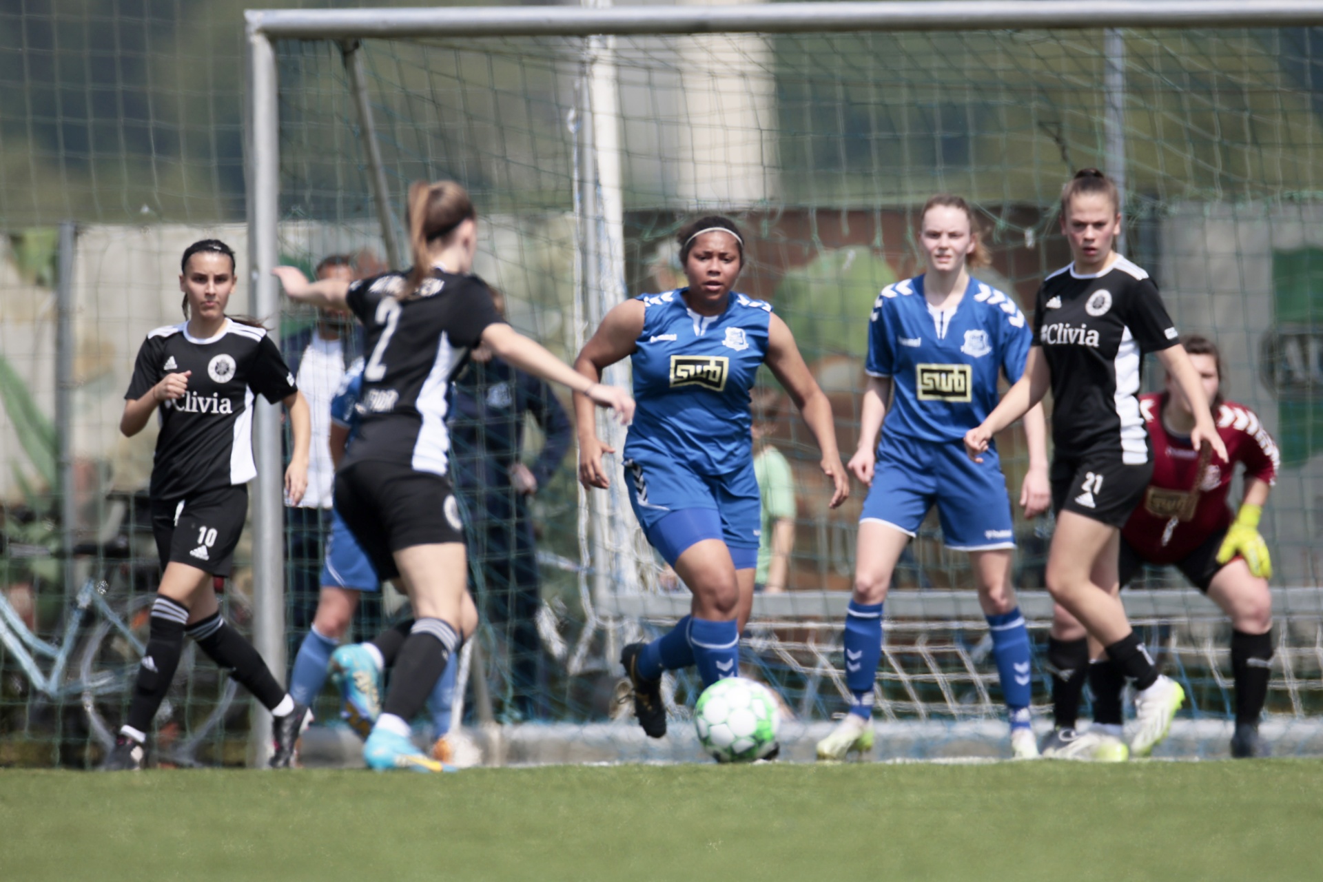 [{"clang_id":1,"value":"Fußballspiel. Frauenmannschaften in Blau und Schwarz-Weiß kämpfen um den Ball auf grünem Rasen vor Zuschauern. Im Hintergrund ein Tornetz."}]