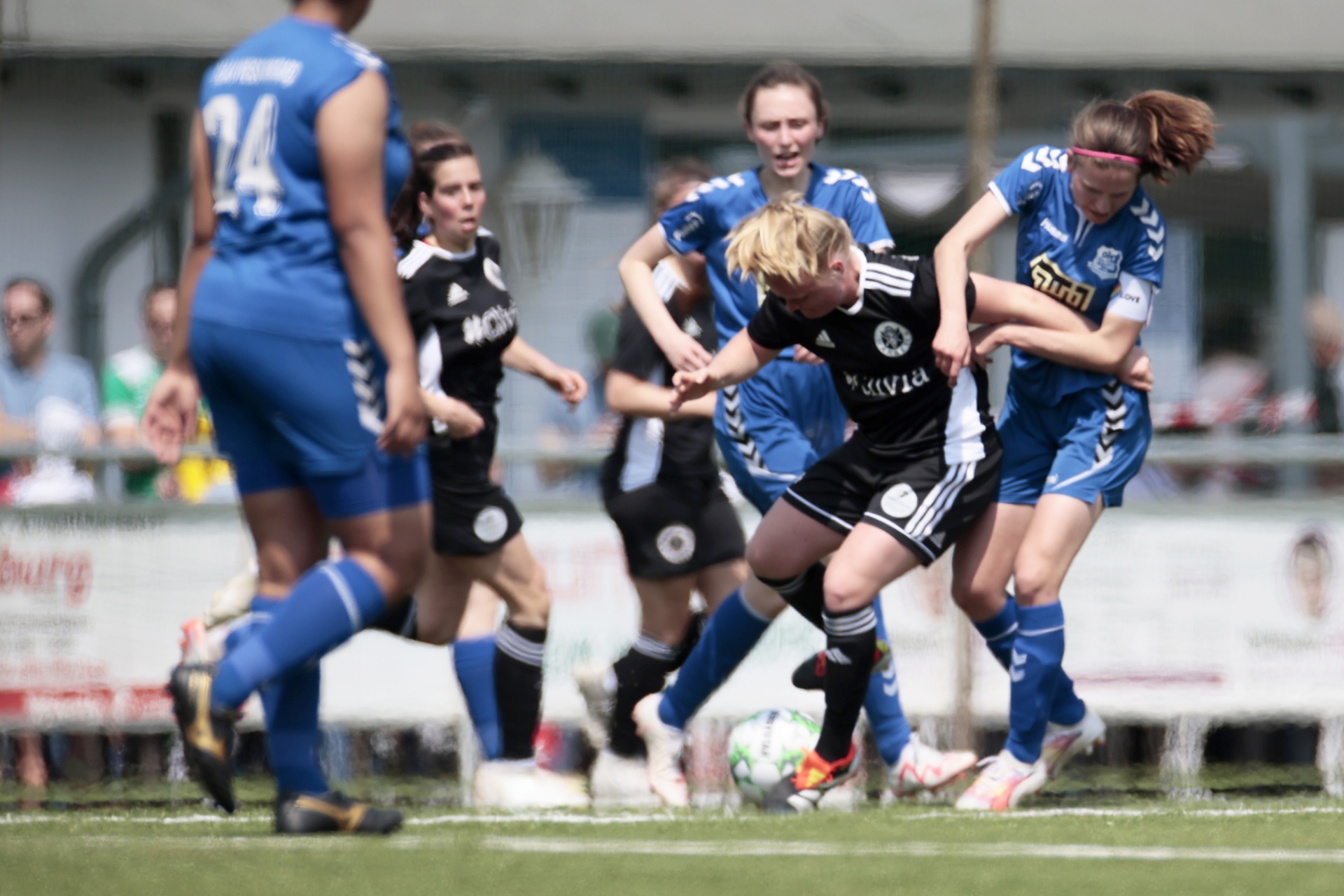 [{"clang_id":1,"value":"Fußballspiel: Spielerinnen kämpfen um den Ball. Zwei in Blau, zwei in Schwarz-Weiß. Im Hintergrund Zuschauer und ein Werbebanner. Rasenplatz."}]