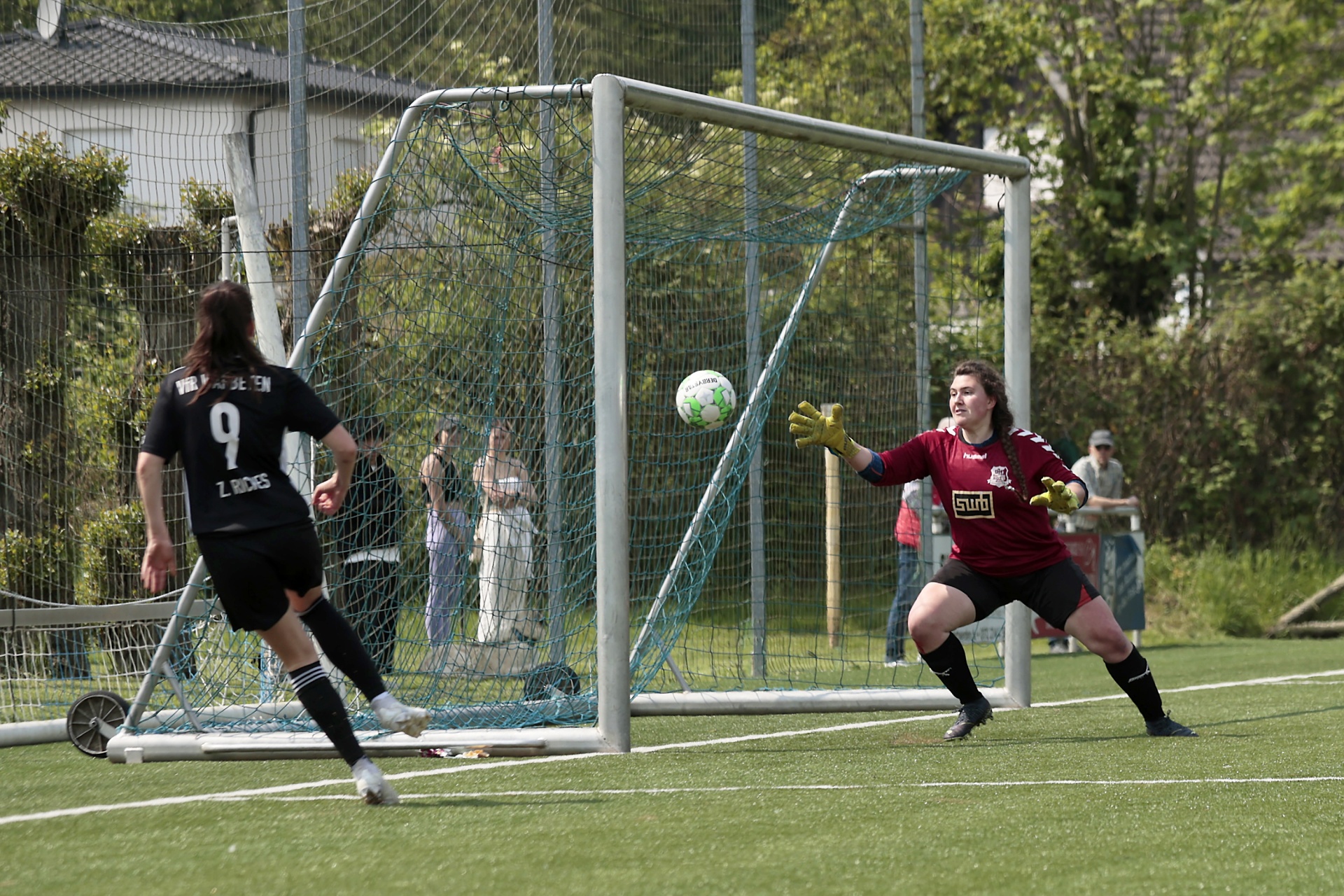 [{"clang_id":1,"value":"Fußballspiel: Spielerin in schwarzem Trikot schießt auf Tor, Torwartin in rot hält den Ball. Im Hintergrund Zuschauer und Bäume."}]