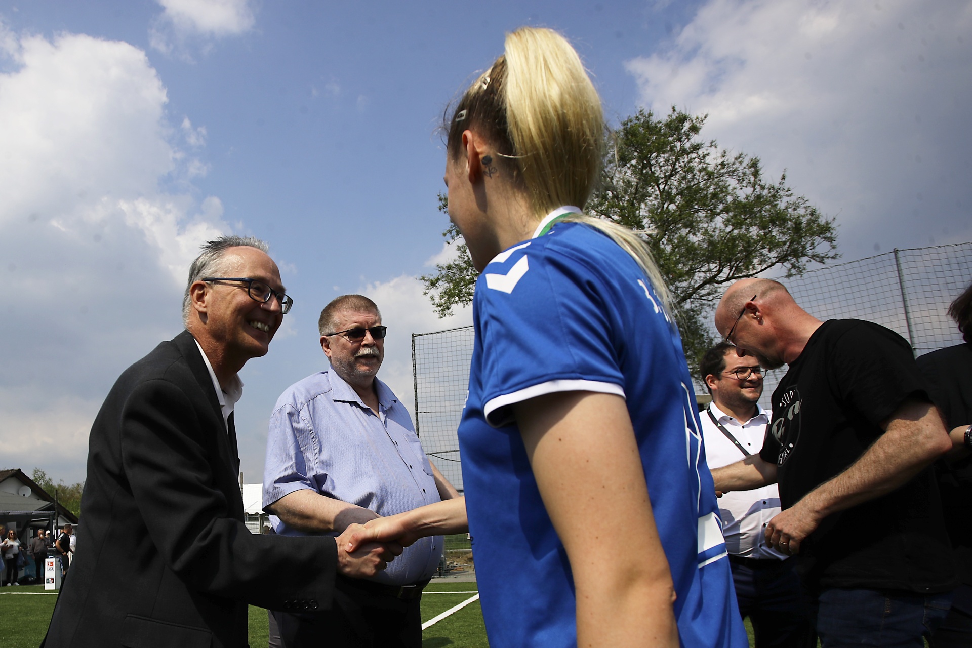 [{"clang_id":1,"value":"Mann im Anzug schüttelt Frau in blauem Sportshirt die Hand. Im Hintergrund weitere Personen und ein Fußballfeld unter blauem Himmel."}]