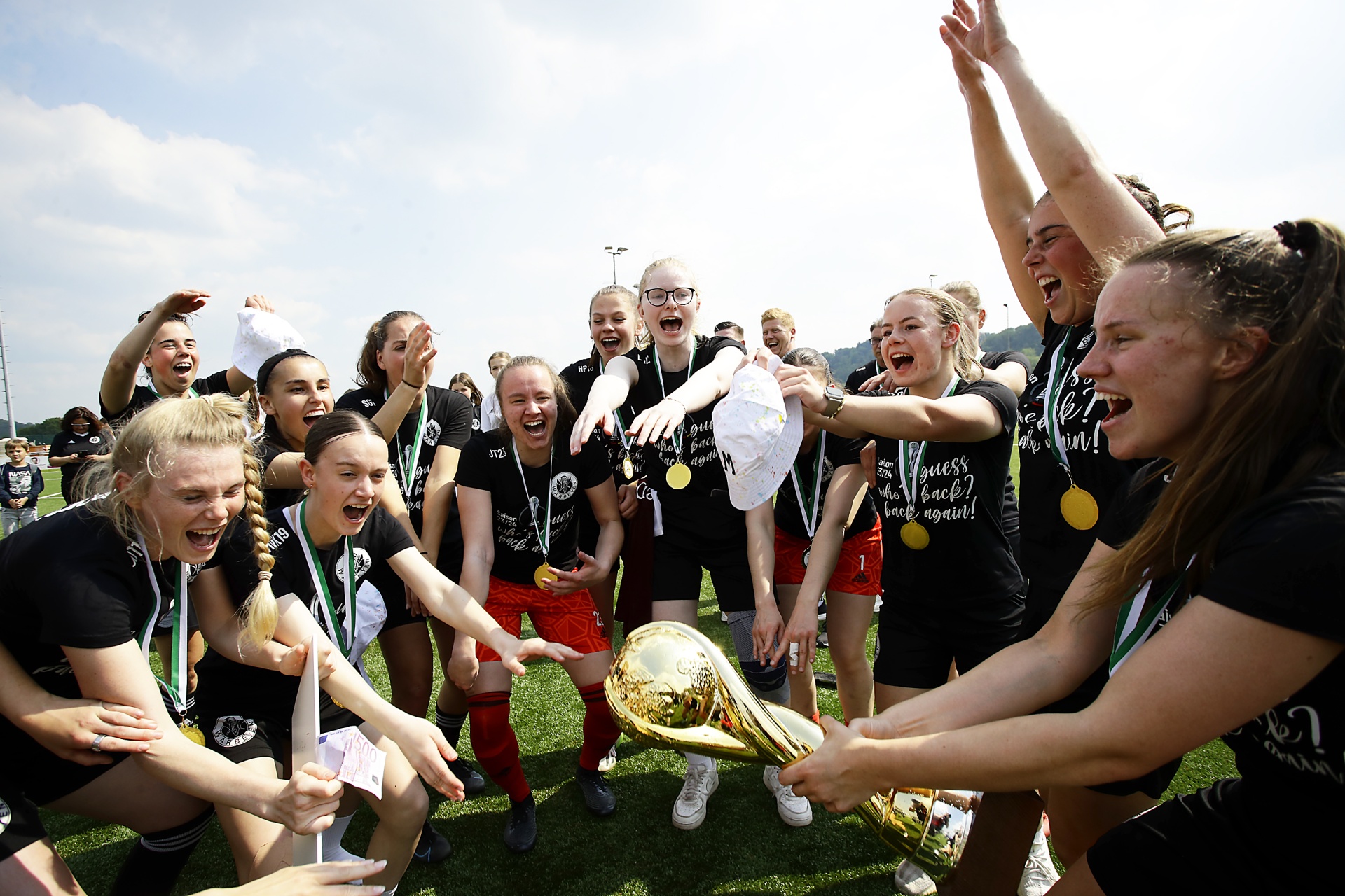 [{"clang_id":1,"value":"Frauenfußballteam jubelt mit Pokal und Medaillen auf einem Rasenplatz. Spielerinnen umarmen sich und heben Arme in die Luft."}]