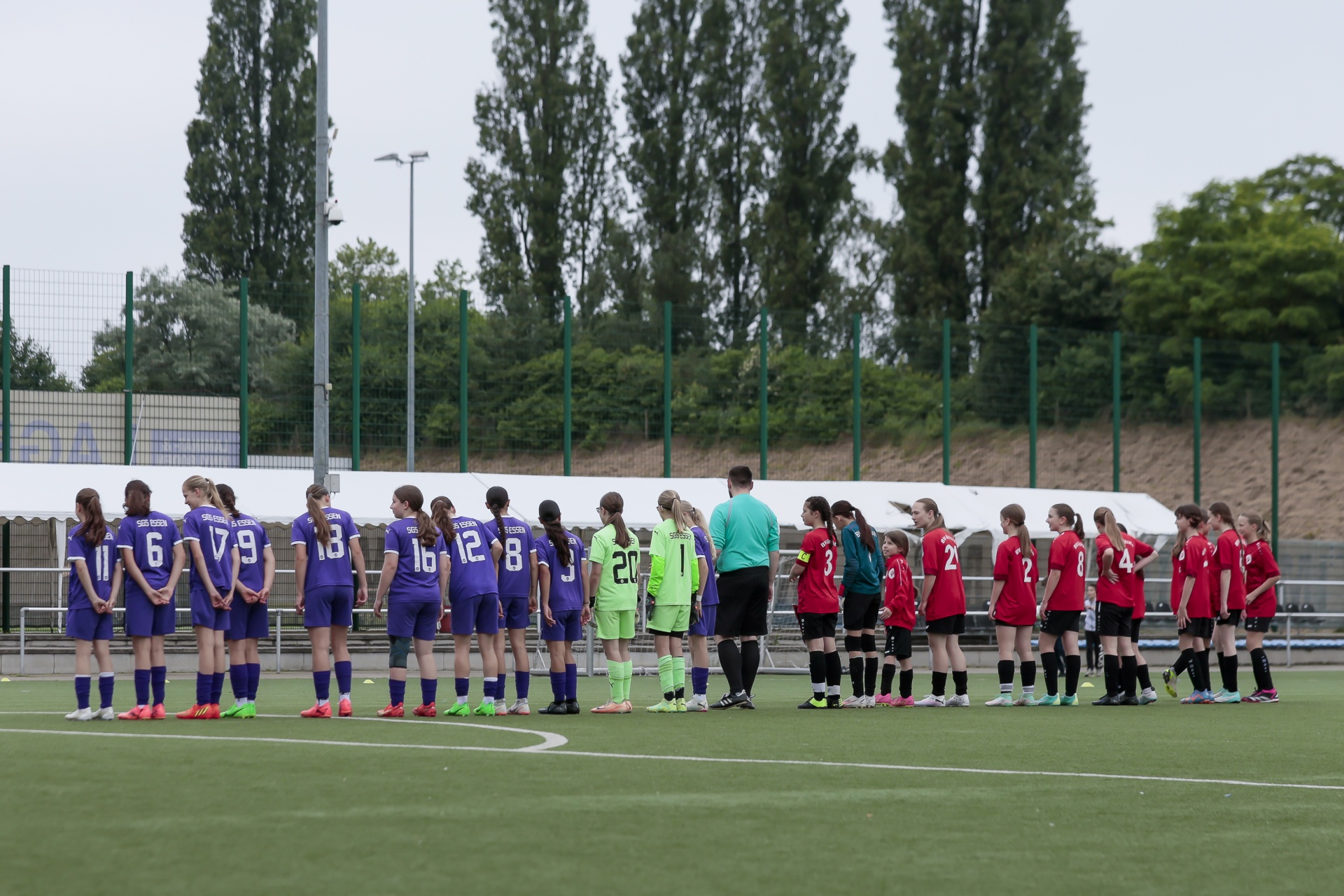[{"clang_id":1,"value":"Fußballspiel: Zwei Jugendmannschaften stehen sich gegenüber. Links in violetten Trikots, rechts in roten. Schiedsrichterin dazwischen. Grünfläche im Hintergrund."}]
