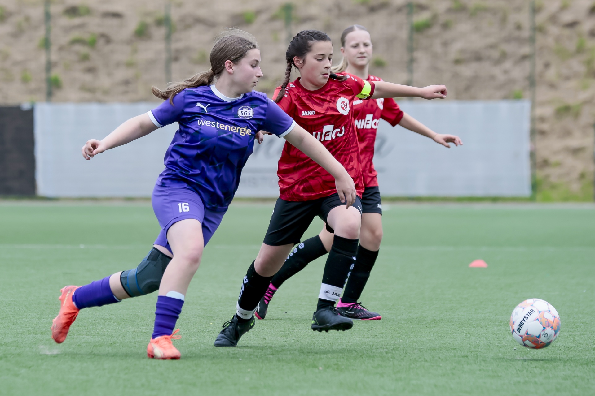 [{"clang_id":1,"value":"Zwei junge Fußballspielerinnen auf grünem Rasen. Links trägt eine Spielerin ein blaues Trikot mit der Nummer 16, rechts ein rotes mit der Aufschrift „Jako“. Eine weitere Spielerin im Hintergrund."}]