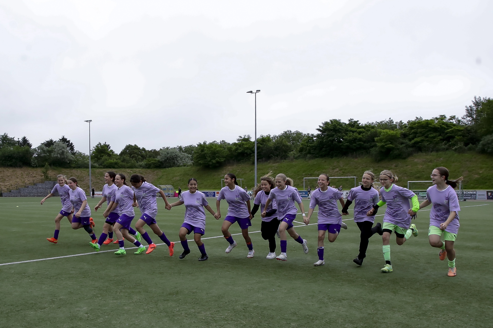 [{"clang_id":1,"value":"Fußballmannschaft aus jungen Frauen in lila Trikots und Shorts läuft auf grünem Kunstrasenfeld. Hügel im Hintergrund, grauer Himmel."}]