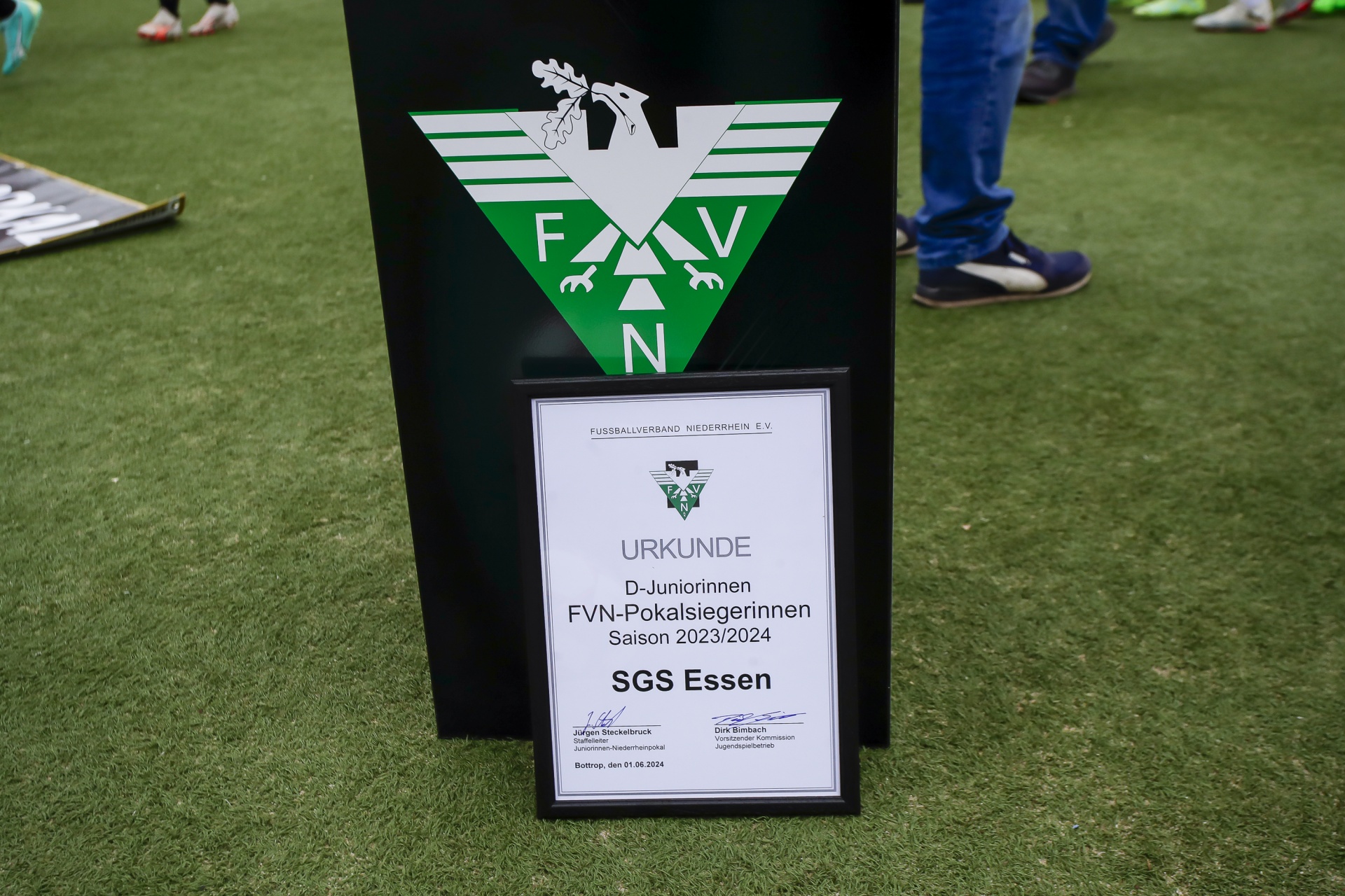 [{"clang_id":1,"value":"Pokalübergabe auf Grün. Im Vordergrund Urkunde mit Schriftzug \"SGS Essen\" und Jahreszahl \"2023/2024\". Im Hintergrund Personen und ein Banner mit Logo \"FVN\"."}]