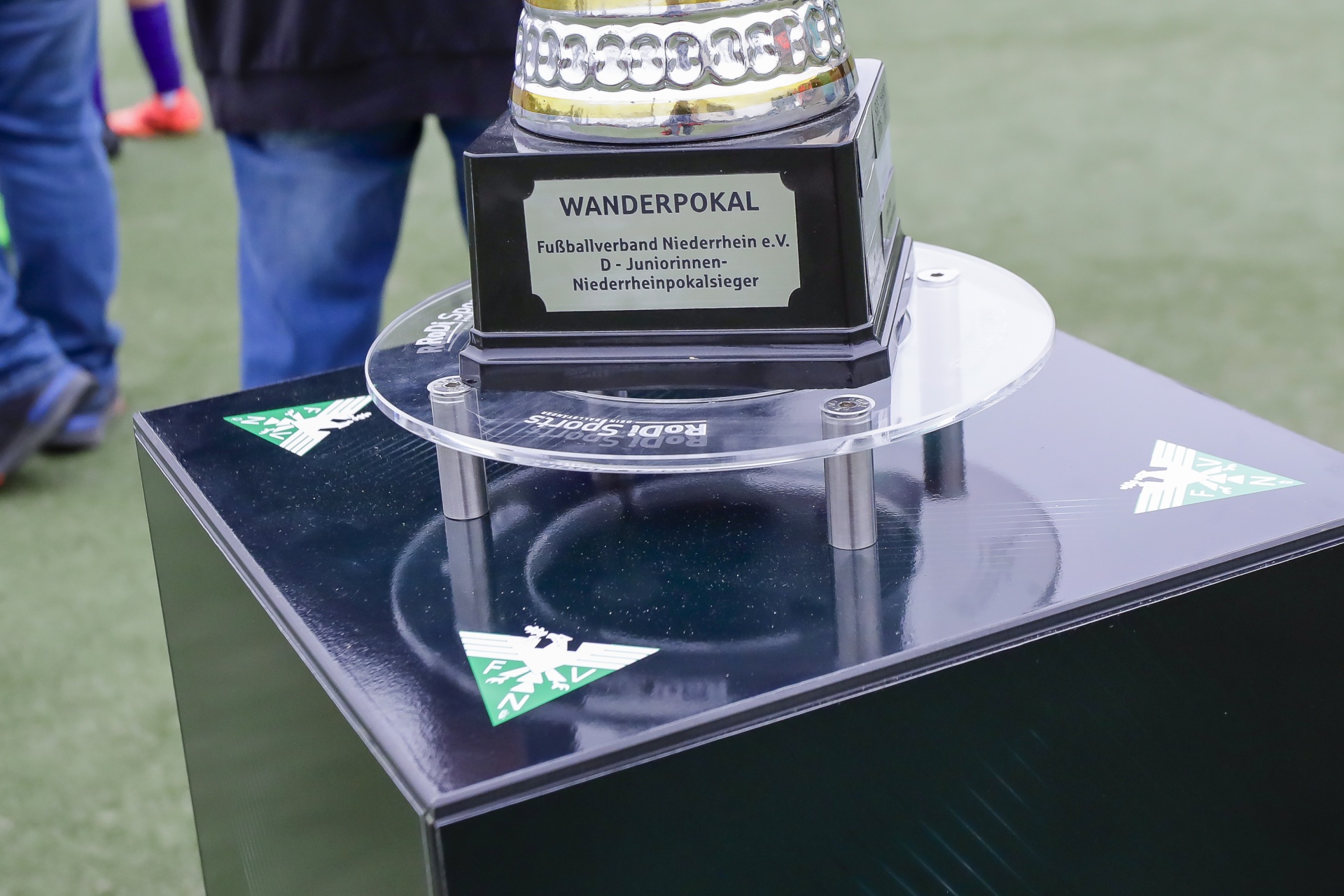 [{"clang_id":1,"value":"Wanderpokal auf schwarzem Podest mit grünem Logo. Beschriftung: \"Fußballverband Niederrhein e.V. D-Juniorinnen-Niederrheinpokalsieger\". Im Hintergrund verschwommen Personen und eine Grünfläche."}]