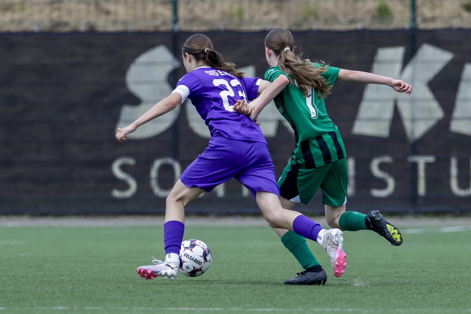 [{"clang_id":1,"value":"Zwei Fußballspielerinnen im Zweikampf. Links trägt eine Spielerin ein violettes Trikot mit der Nummer 23, rechts ein grün-schwarzes. Im Hintergrund eine graue Wand mit Schriftzug."}]