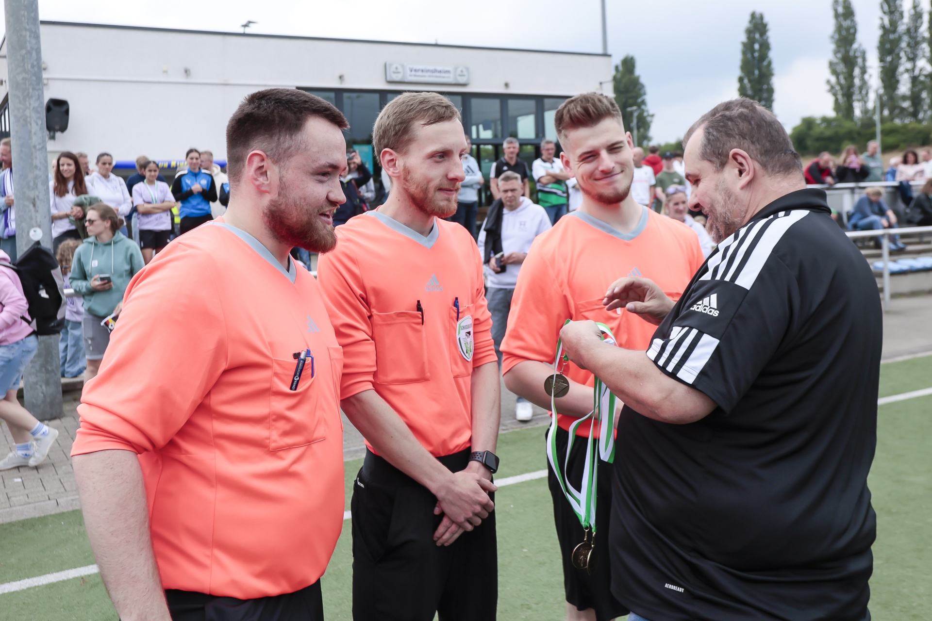 [{"clang_id":1,"value":"Drei Fußballschiedsrichter in orangefarbenen Trikots stehen an der Seitenlinie eines Fußballfeldes. Ein Schiedsrichter in schwarz-weißem Trikot hängt ihnen Medaillen um den Hals. Zuschauer im Hintergrund."}]