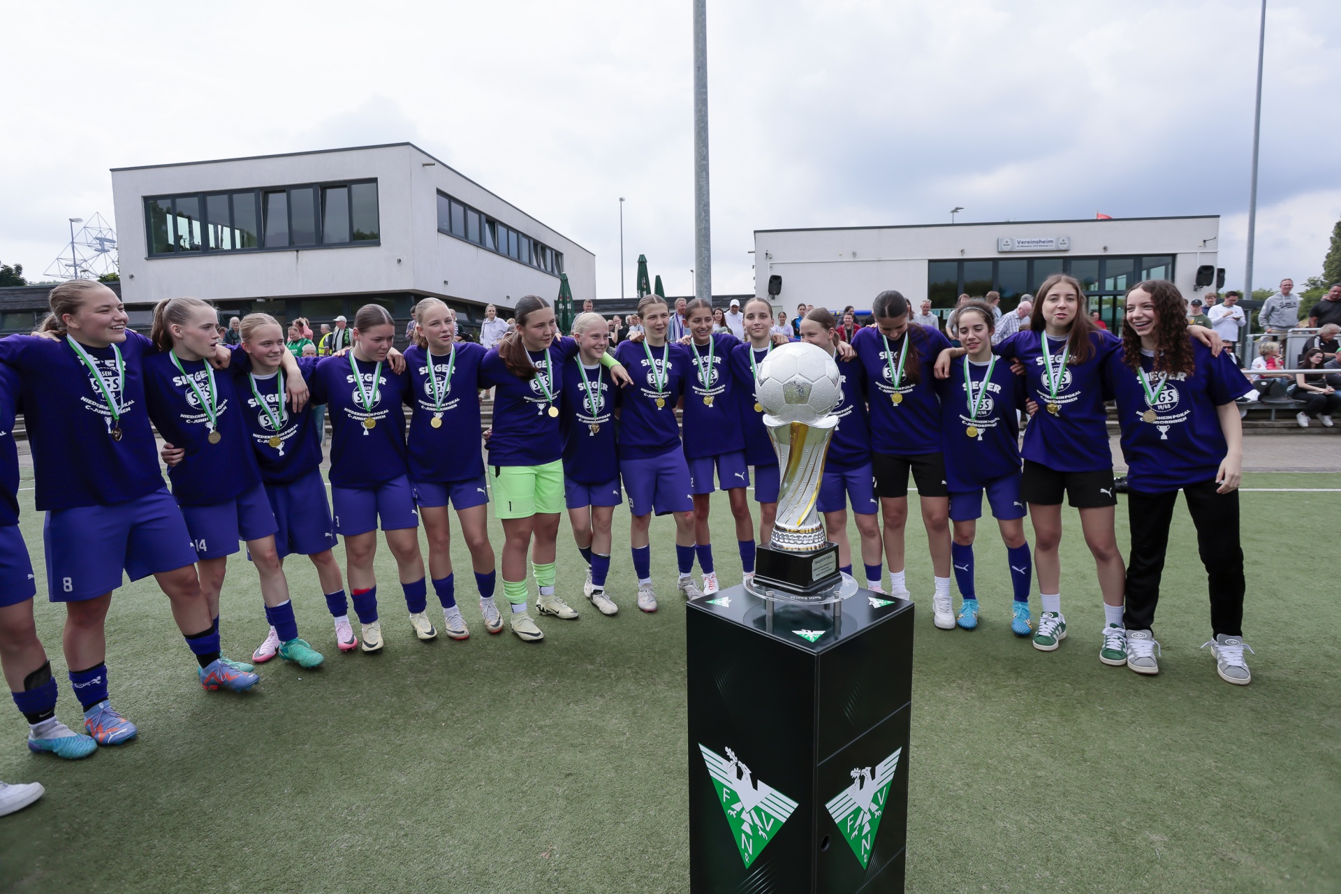 [{"clang_id":1,"value":"Fußballmannschaft mit Medaillen und Pokal auf grünem Kunstrasen. Im Hintergrund Gebäude und Zuschauer."}]