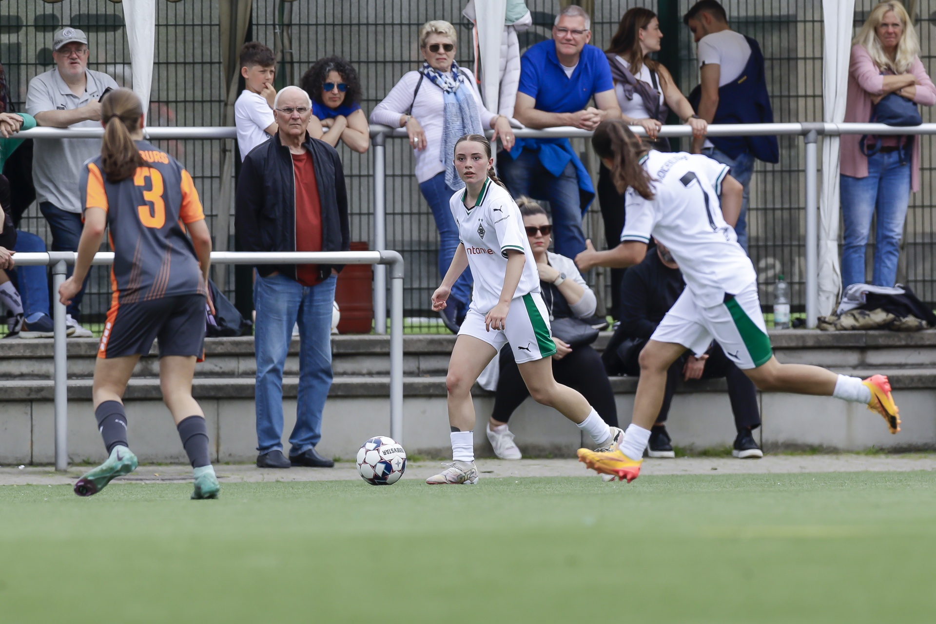 [{"clang_id":1,"value":"Fußballspiel: Zwei Spielerinnen in weißen Trikots mit grünem Saum und eine Spielerin mit orange-braunen Trikot rennen auf einem grünen Rasenfeld. Im Hintergrund Zuschauer auf einer Tribüne."}]