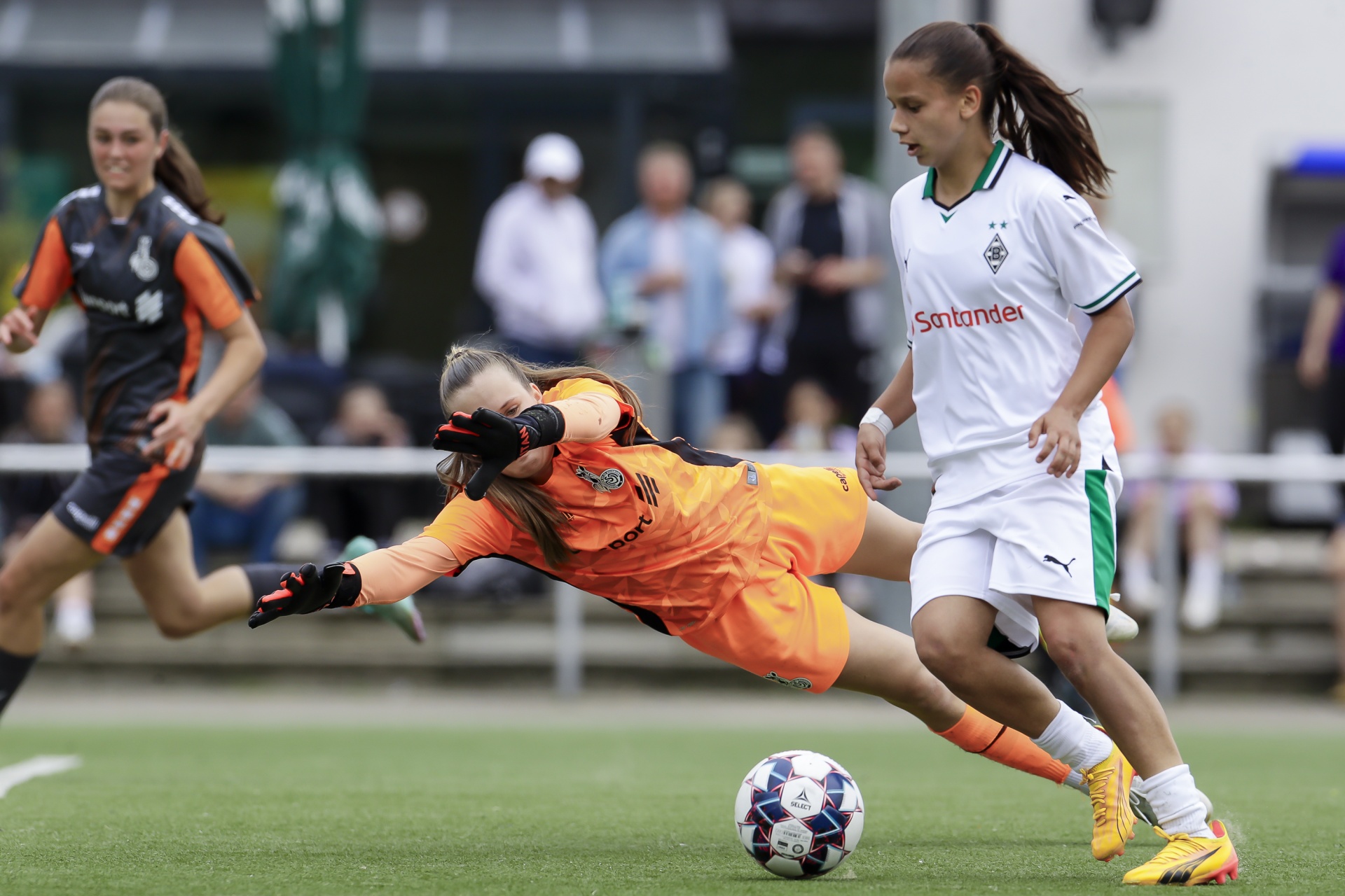 [{"clang_id":1,"value":"Fußballspiel: Torwartin in orange-schwarzem Trikot grätscht auf Ball zu, während Spielerin in weiß-grünem Trikot an ihr vorbeiläuft. Im Hintergrund Zuschauer."}]