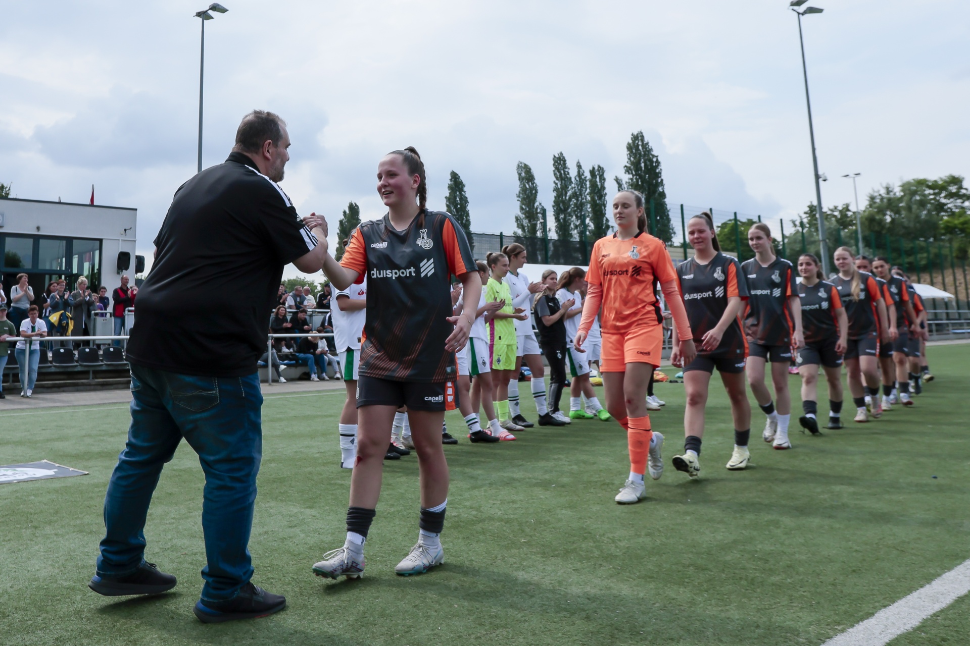 [{"clang_id":1,"value":"Fußballspielerinnen und ein Mann stehen auf einem Fußballfeld. Einige Spielerinnen tragen schwarze Trikots mit \"duisport\"-Logo, andere orange Trikots. Im Hintergrund Zuschauer."}]