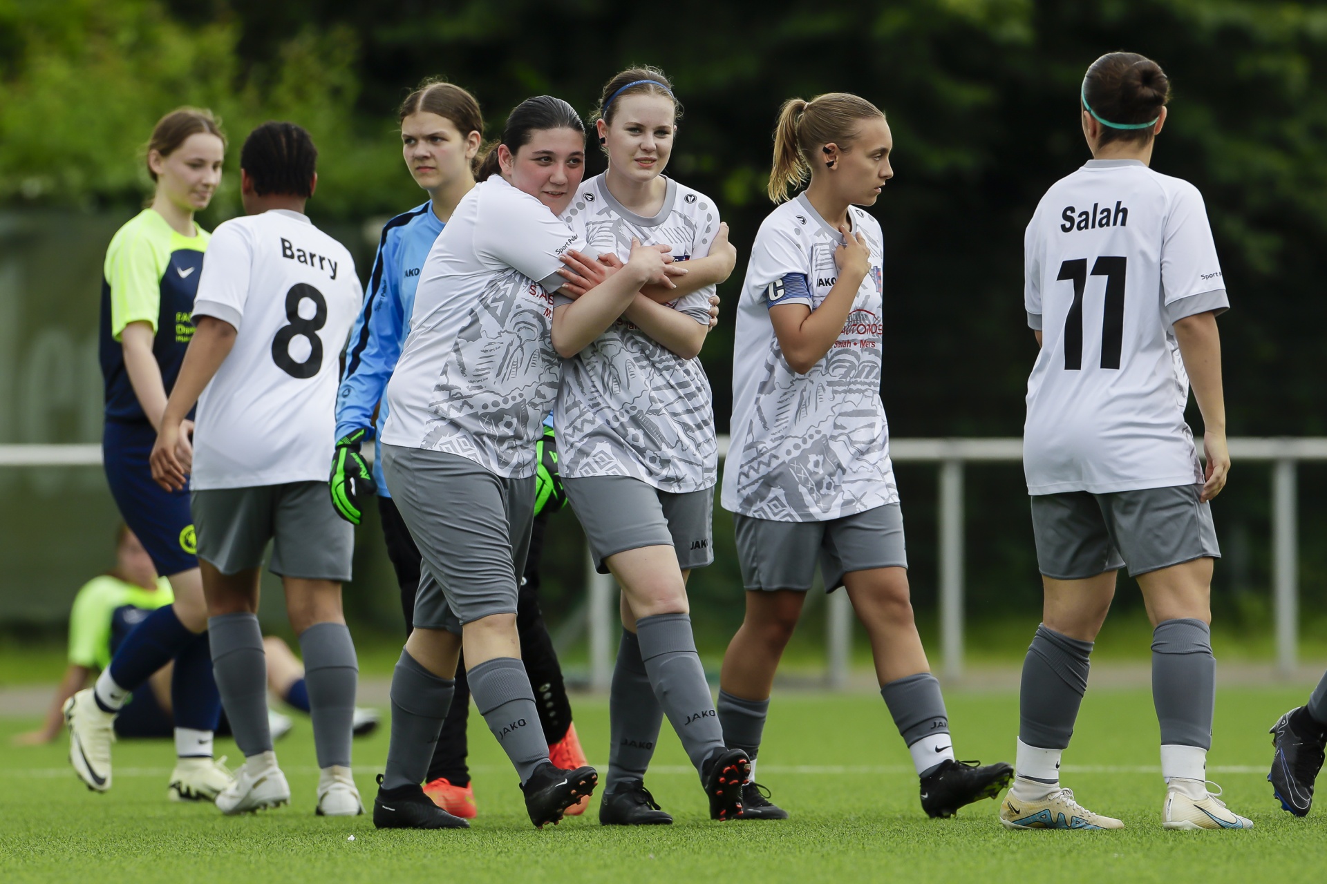 [{"clang_id":1,"value":"Fußballmannschaft junger Frauen umarmt sich auf grünem Rasen. Spieler tragen helle Trikots mit Nummern und graue Shorts. Hintergrund: Bäume und andere Spieler."}]