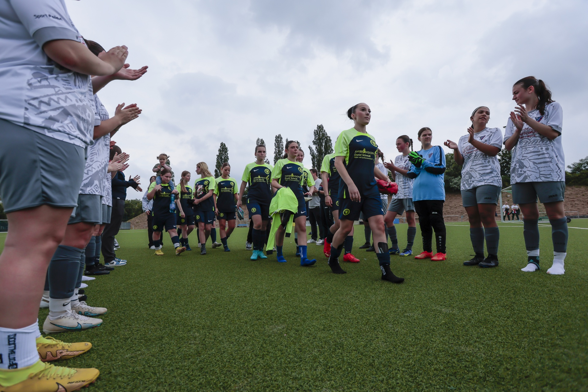 [{"clang_id":1,"value":"Fußballspielerinnen stehen in Formation auf einem grünen Rasen und werden von einer Gruppe Personen geklatscht. Einige Spielerinnen tragen grüne Trikots."}]