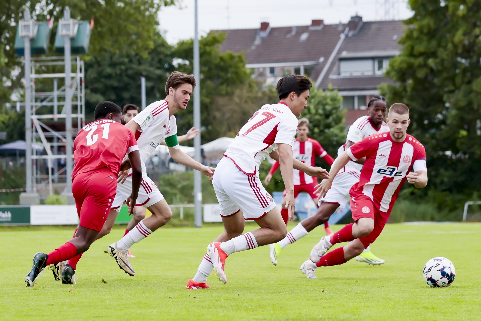 [{"clang_id":1,"value":"Fußballspiel: Mehrere Spieler in roten Trikots laufen auf einem grünen Rasenfeld. Ein Fußball liegt im Vordergrund. Im Hintergrund Gebäude und Bäume."}]