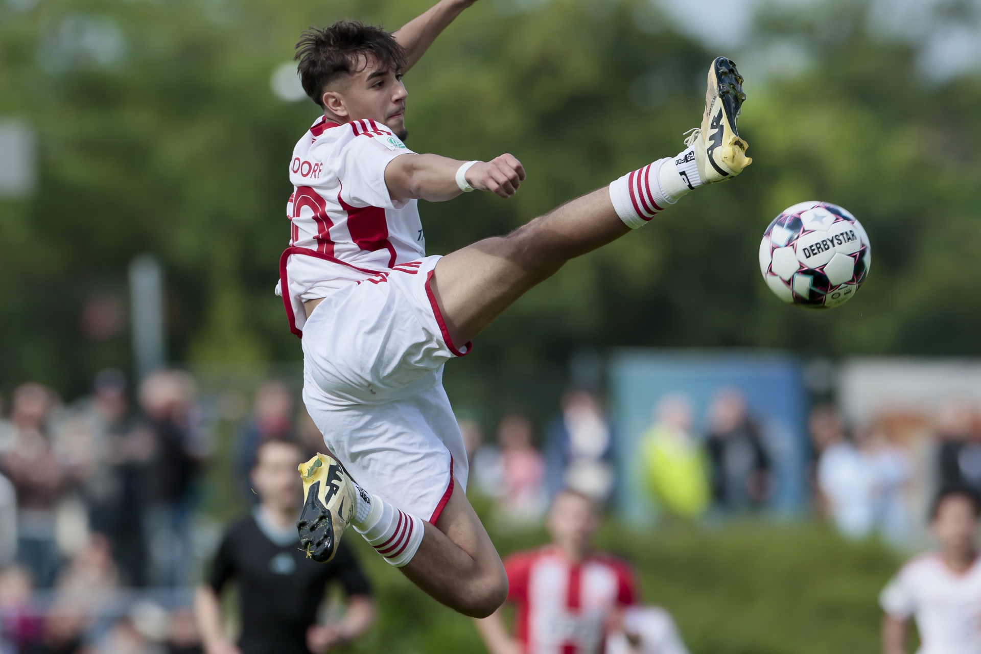 [{"clang_id":1,"value":"Fußballspieler in Rot-Weiß springt mit gestrecktem Bein, um den Ball zu treten. Hintergrund: verschwommene Zuschauer und Grünfläche."}]