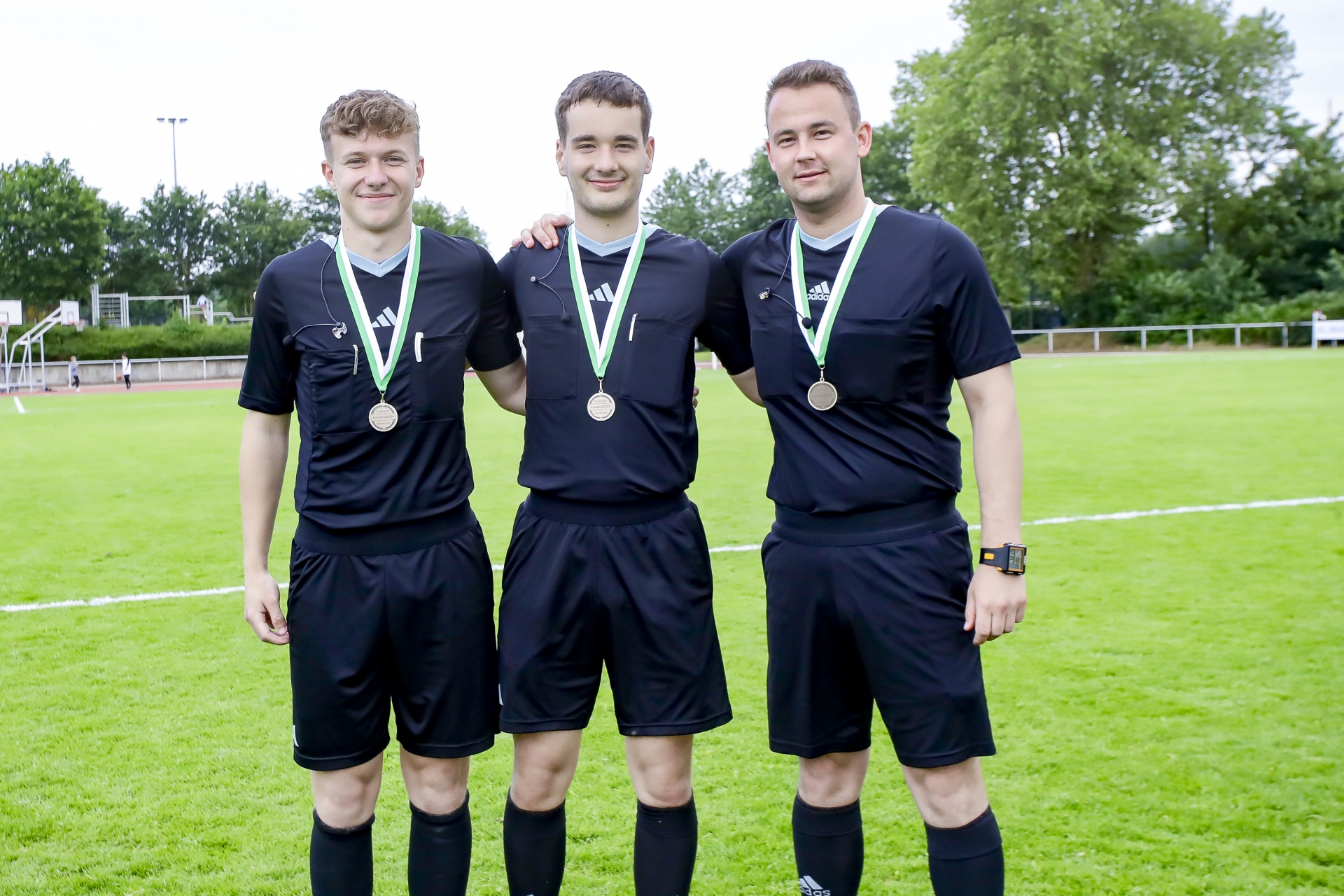 [{"clang_id":1,"value":"Drei junge Männer in schwarzer Fußballschiedsrichterausrüstung mit Pfeifen und Medaillen stehen auf einem Rasenfeld. Im Hintergrund Bäume und ein Sportplatz."}]