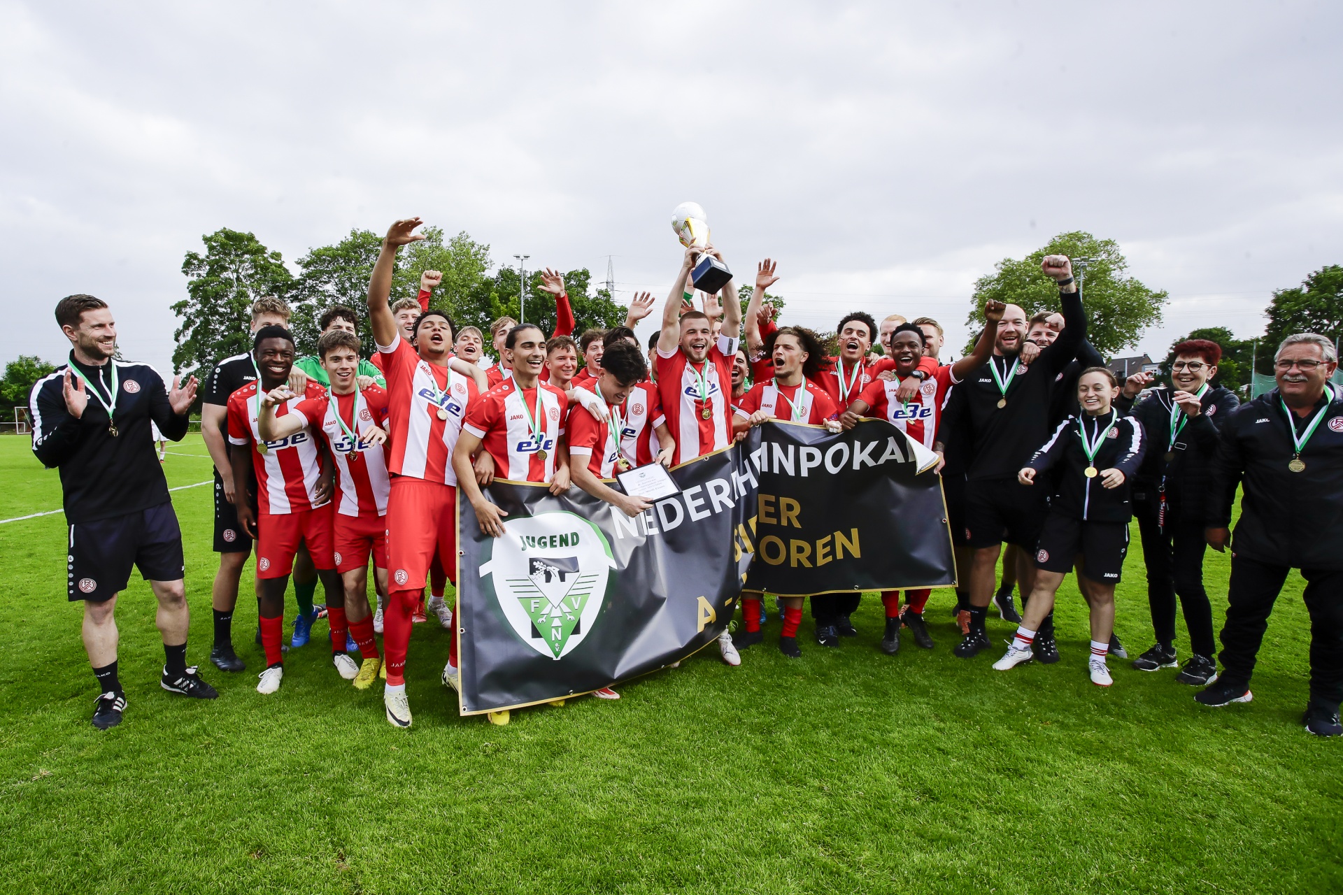 [{"clang_id":1,"value":"Fußballmannschaft in rot-weiß gestreiften Trikots feiert Sieg mit Pokal und Banner auf grünem Rasen. Zuschauer im Hintergrund."}]