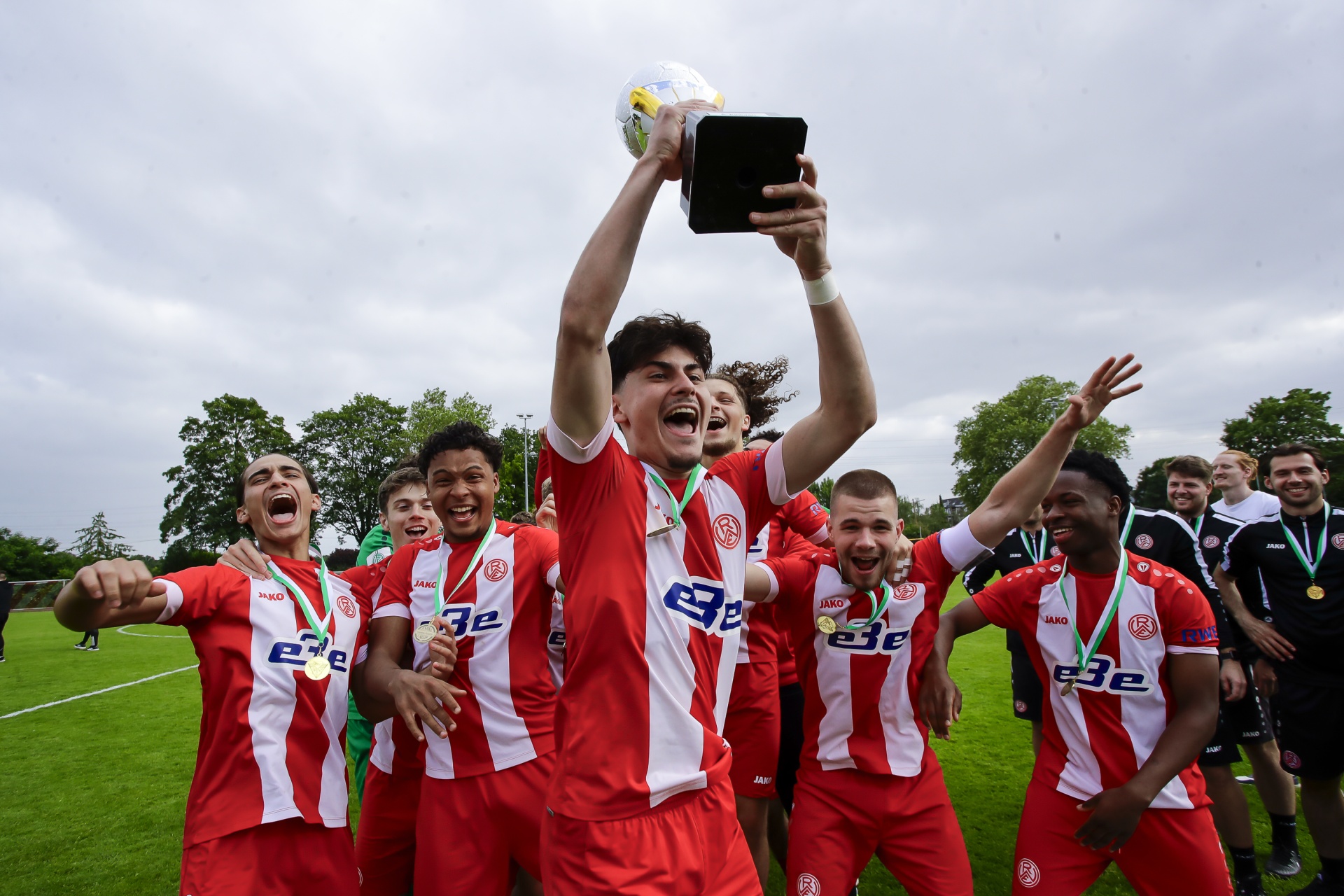 [{"clang_id":1,"value":"Fußballmannschaft in rot-weißen Trikots feiert Sieg auf Rasen. Spieler halten Pokal und Smartphone hoch, jubeln und tragen Medaillen."}]