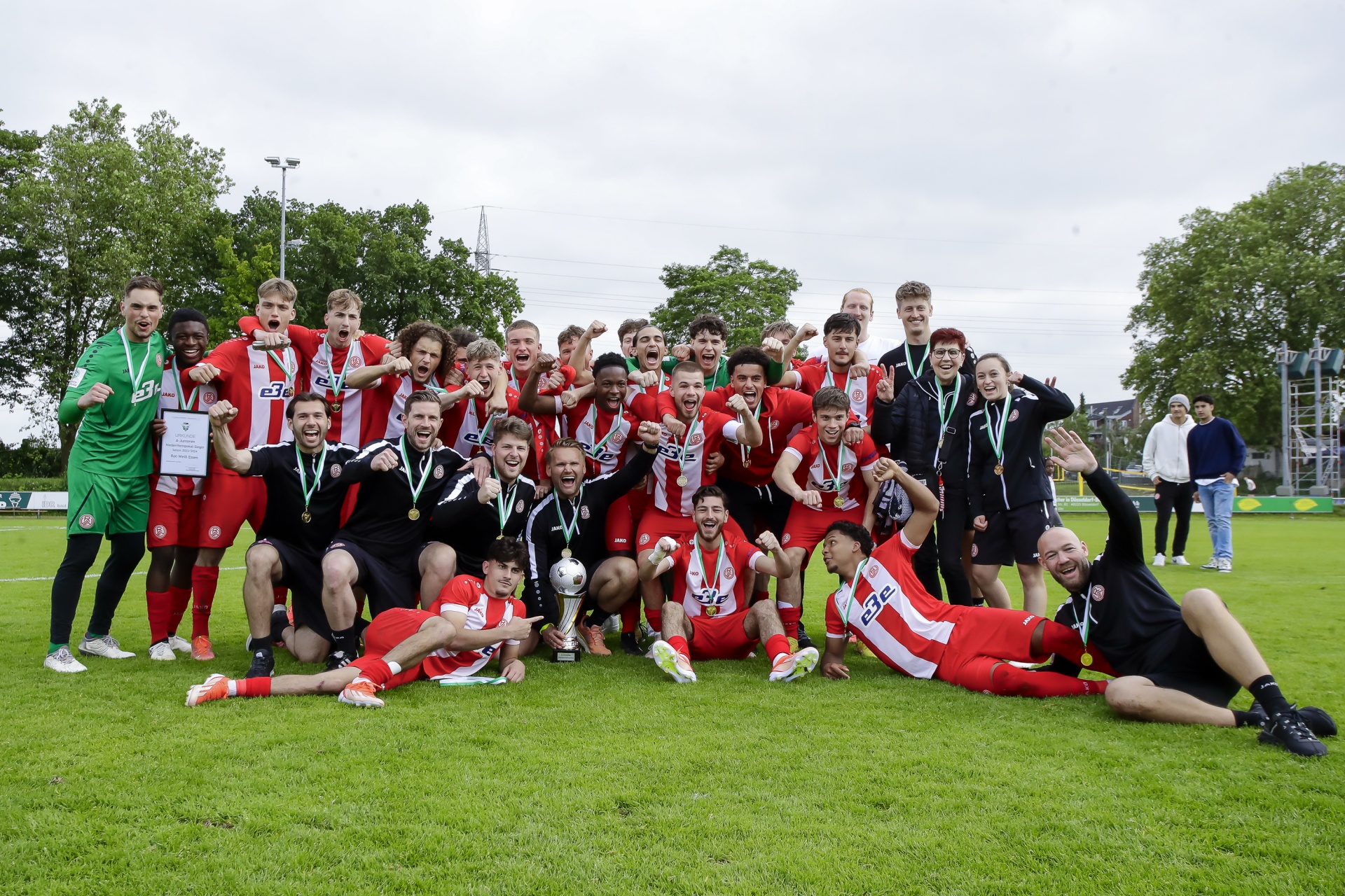 [{"clang_id":1,"value":"Fußballmannschaft in Rot-Weiß feiert Sieg auf Grünfläche. Spieler mit Medaillen und Pokal posieren strahlend, einige liegen lachend im Gras. Im Hintergrund Zuschauer und Gebäude."}]