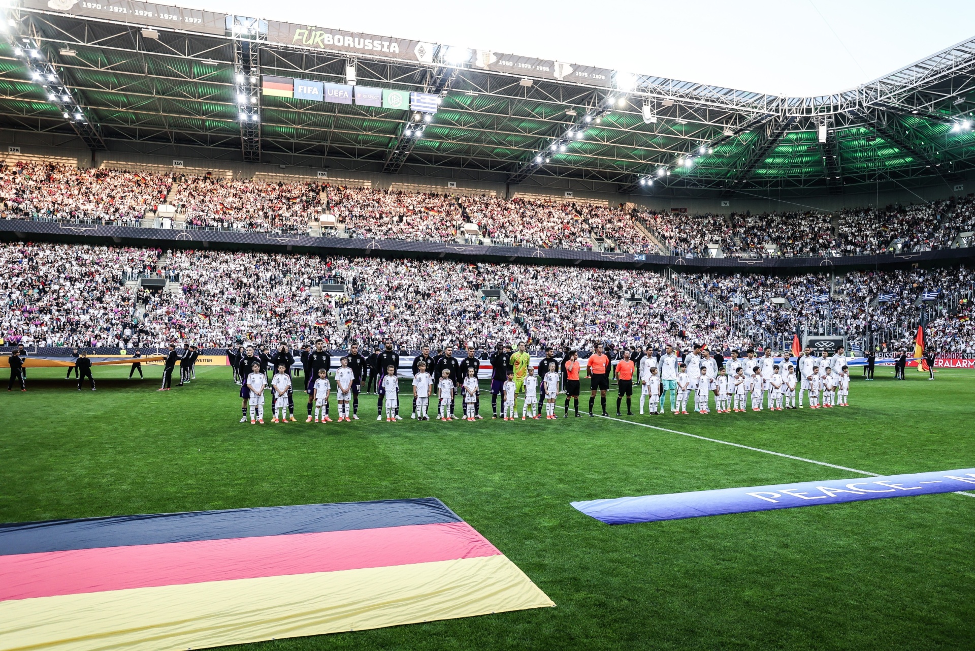 [{"clang_id":1,"value":"Fußballstadion gefüllt mit Zuschauern. Spieler stehen in zwei Reihen auf dem Rasen, Arm in Arm. Im Vordergrund liegen Flaggen mit den Farben Deutschland, Rot, Schwarz und Gold."}]