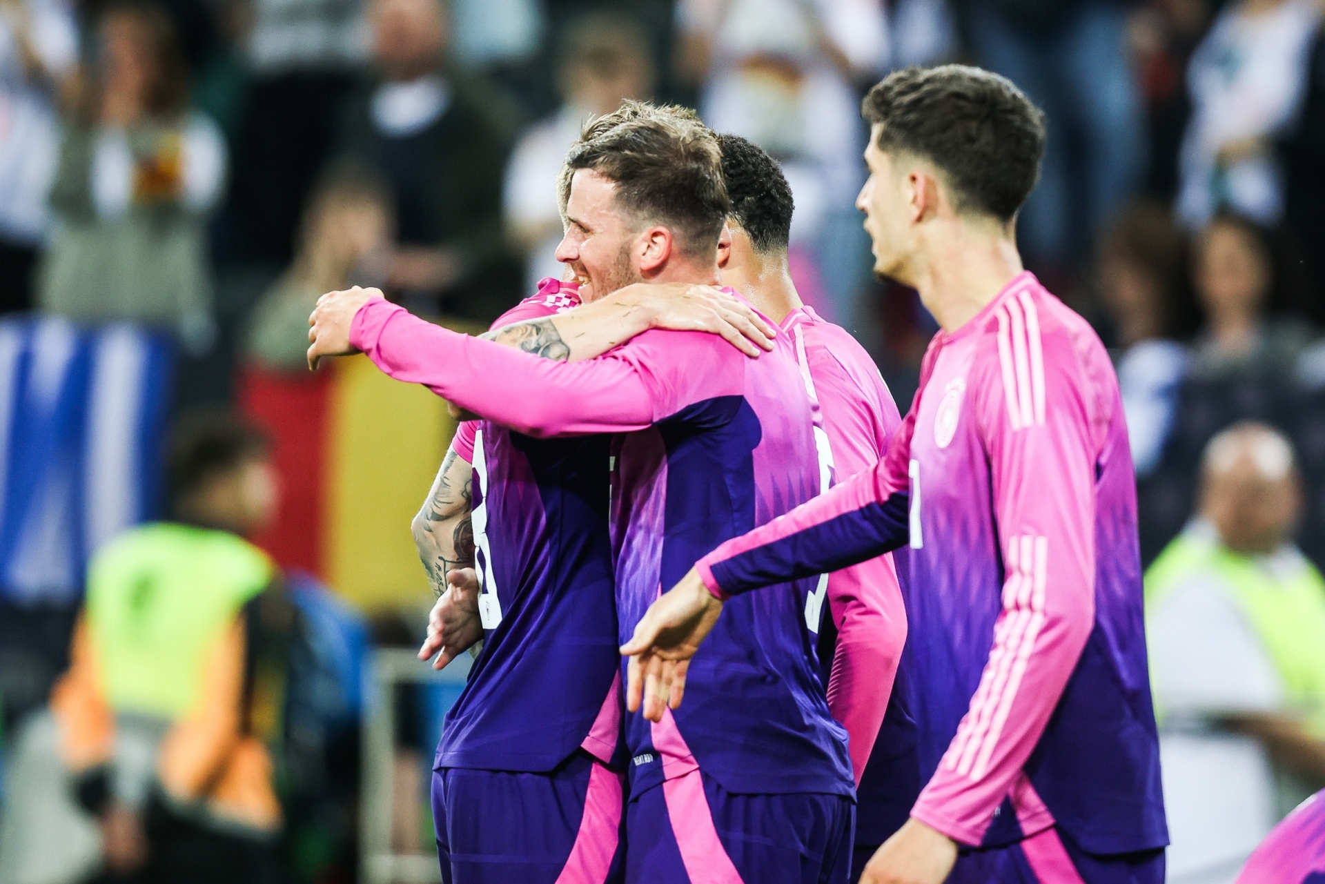 [{"clang_id":1,"value":"Drei Fußballspieler im Vereinstrikot (lila-pink) feiern einen Spielzug mit eng umschlungenen Armen. Im Hintergrund verschwommene Zuschauer."}]